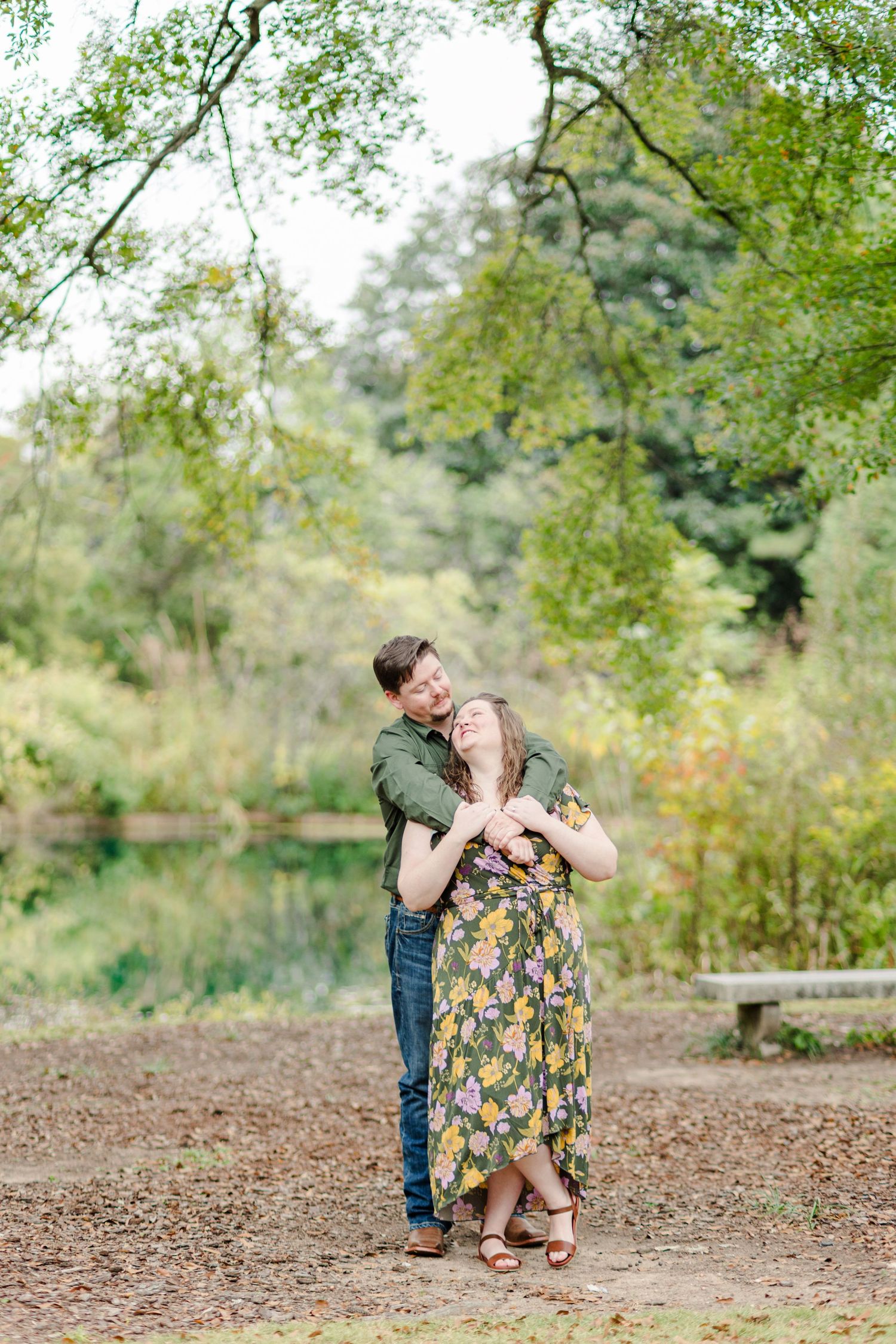 Couple embracing in a lush green park, woman in floral dress laughing joyfully, man hugging her from behind near a pond.