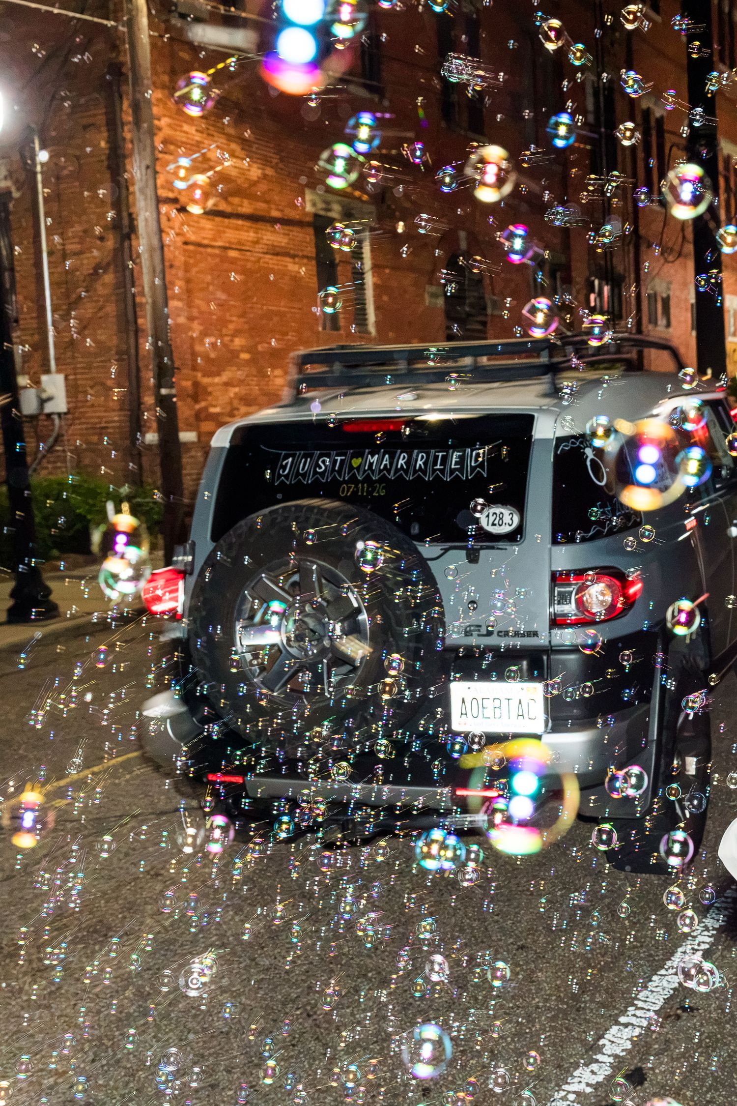 Just Married SUV surrounded by colorful soap bubbles outside brick building at night wedding sendoff.