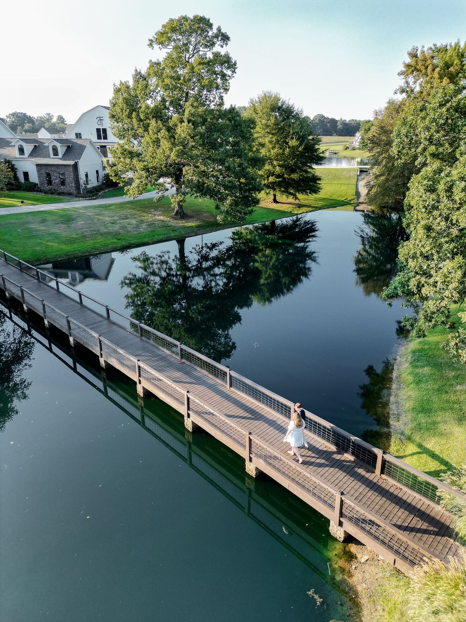 Aerial view of a bride on a wooden bridge over a calm river, surrounded by lush trees and a white farmhouse venue.
