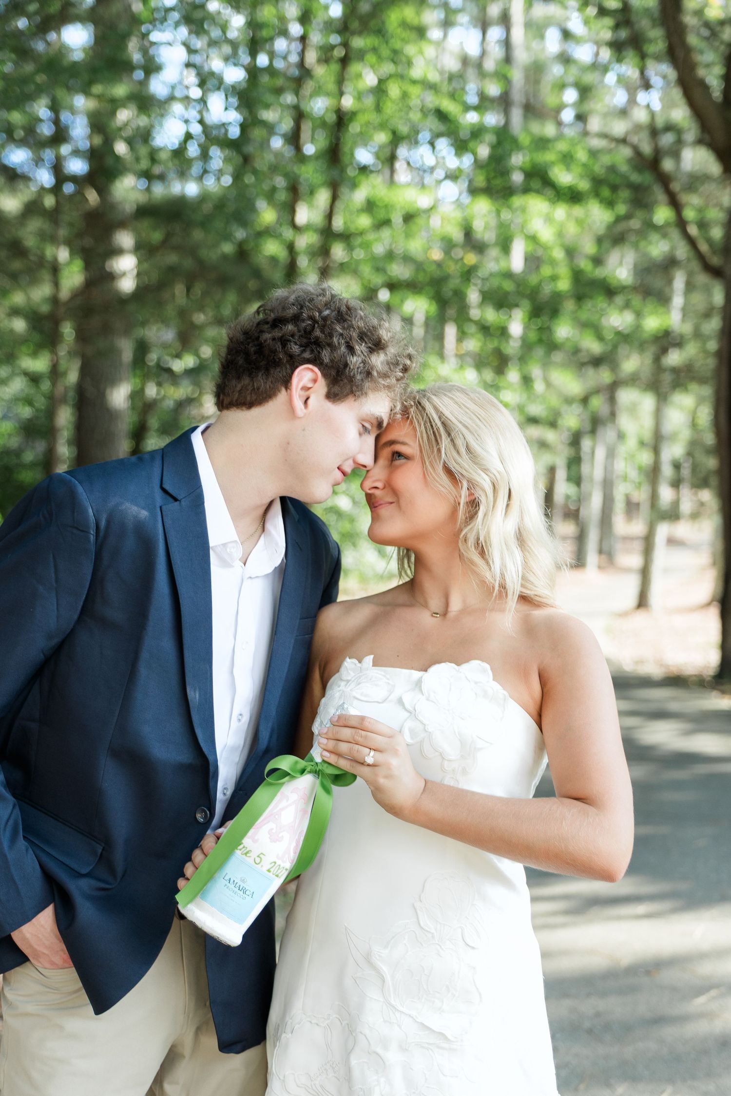 Bride and groom in navy suit share romantic moment holding bottle in sunlit pine forest.