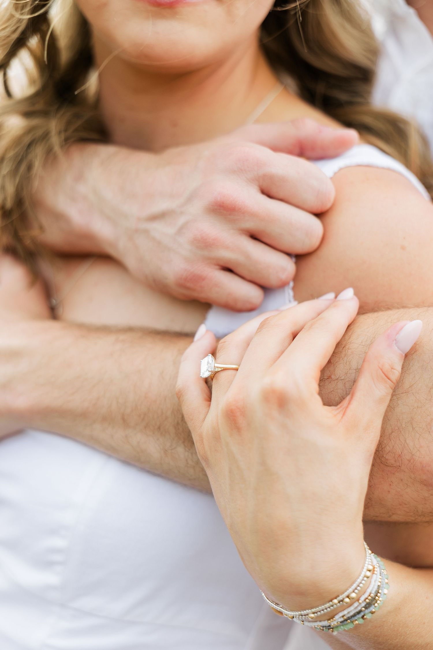 Close-up of couple embracing, woman displaying gold diamond engagement ring and beaded bracelets on man's arm.