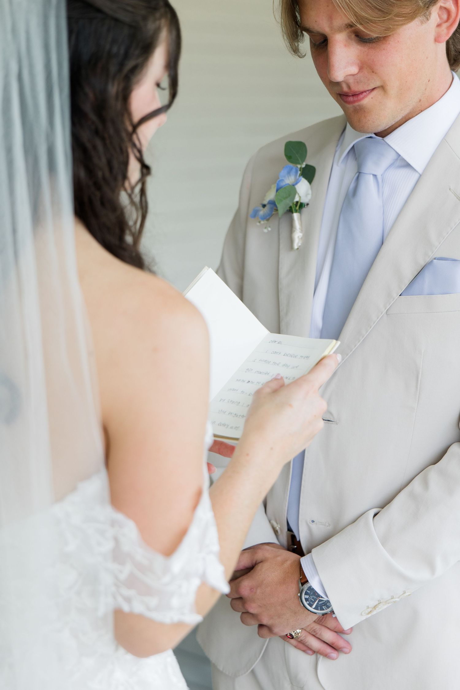 Bride reads wedding vows to groom in tan suit with blue floral boutonniere during outdoor ceremony.
