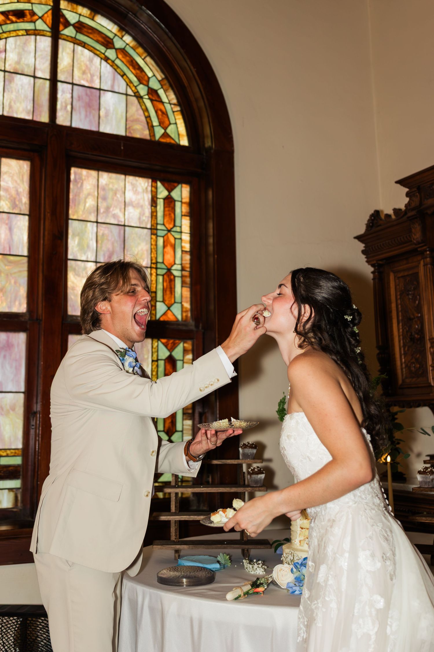 Bride and groom share a playful wedding cake feeding moment in front of colorful stained glass window.