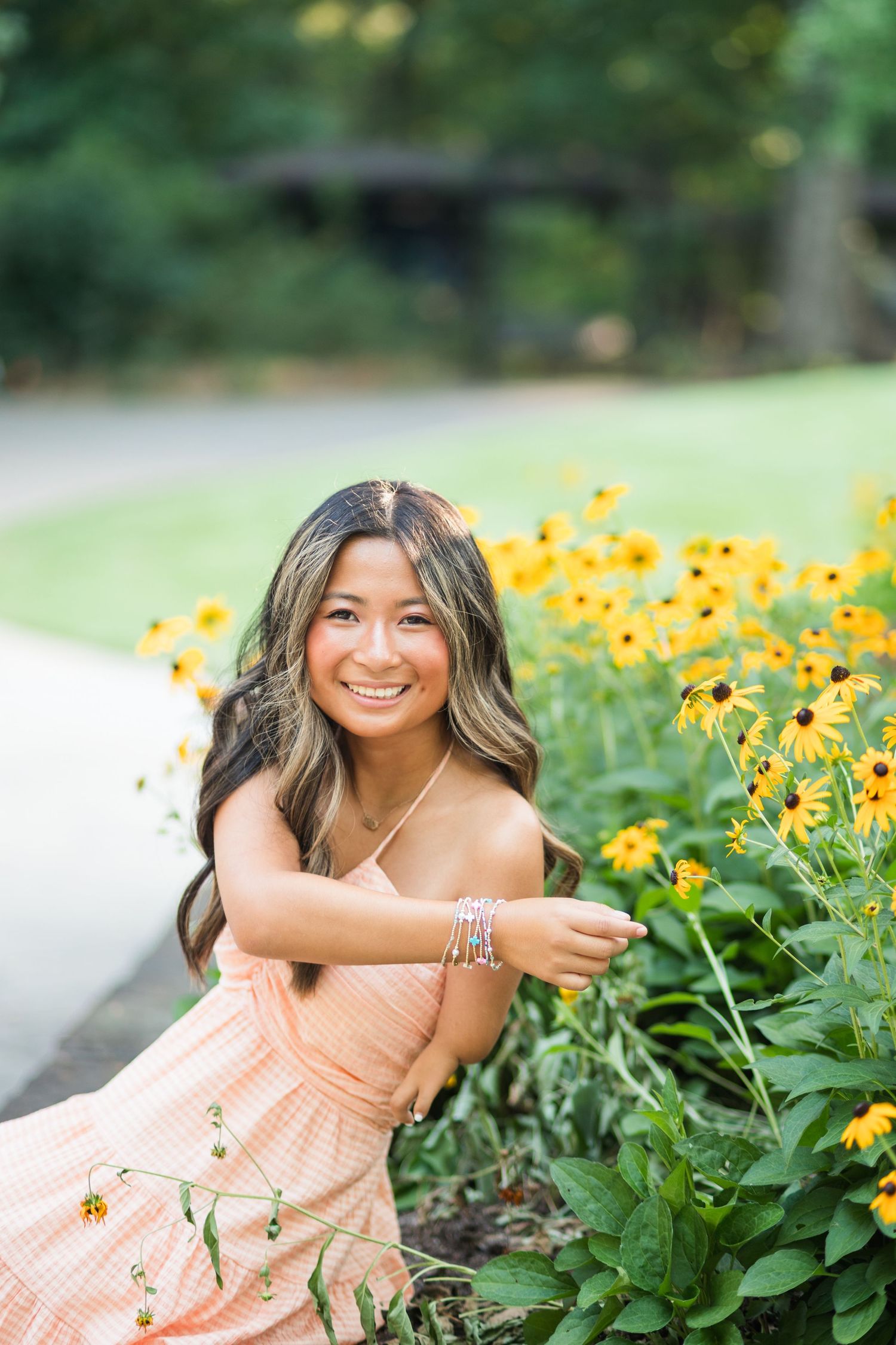 Smiling young woman in peach dress crouching beside yellow black-eyed Susan flowers.