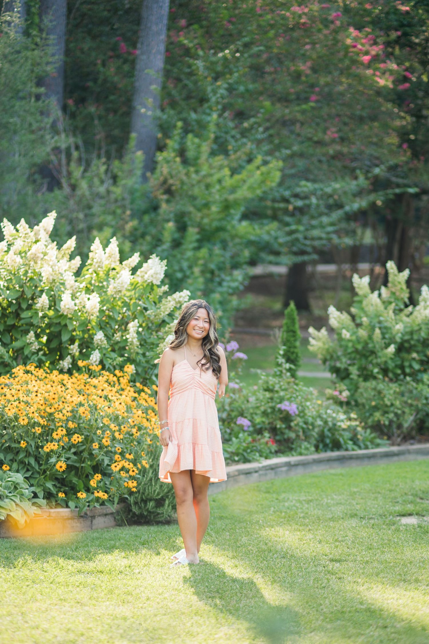 Young woman in peach dress strolling through colorful garden with yellow and white flowers.