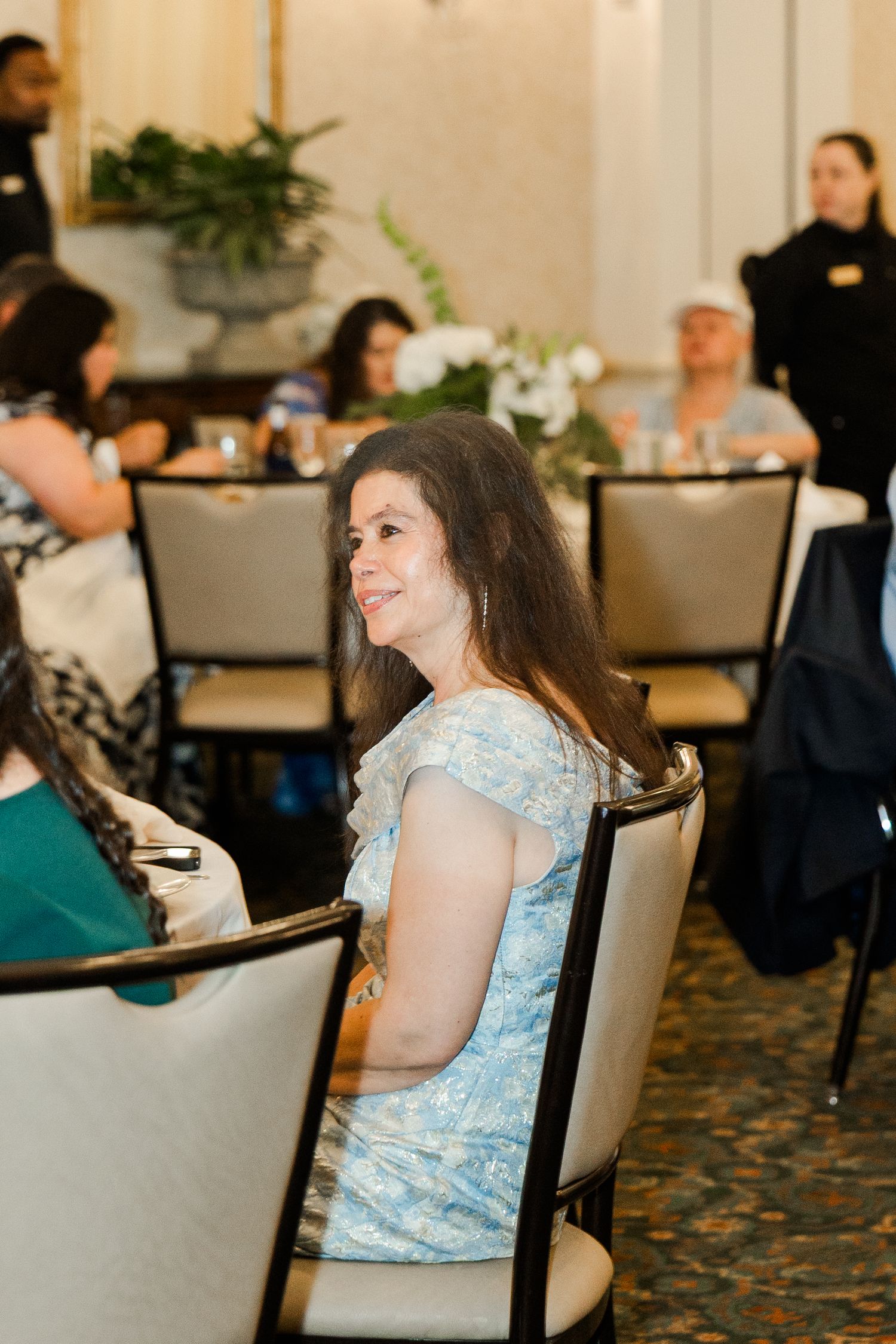 Smiling female wedding guest in blue floral dress seated at elegantly decorated reception dinner table.