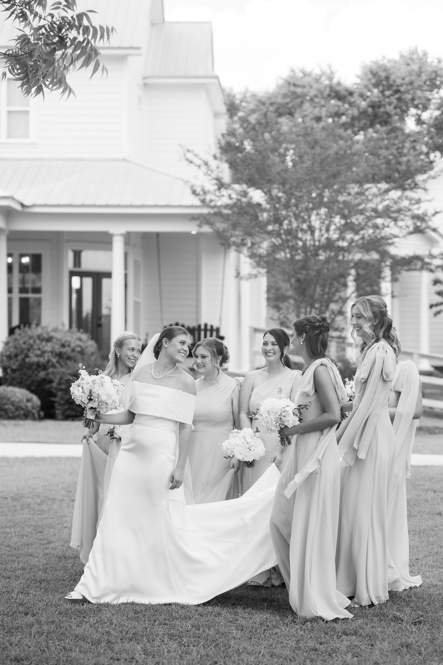 Black and white photo of bride and bridesmaids gathered on lawn in front of white farmhouse.
