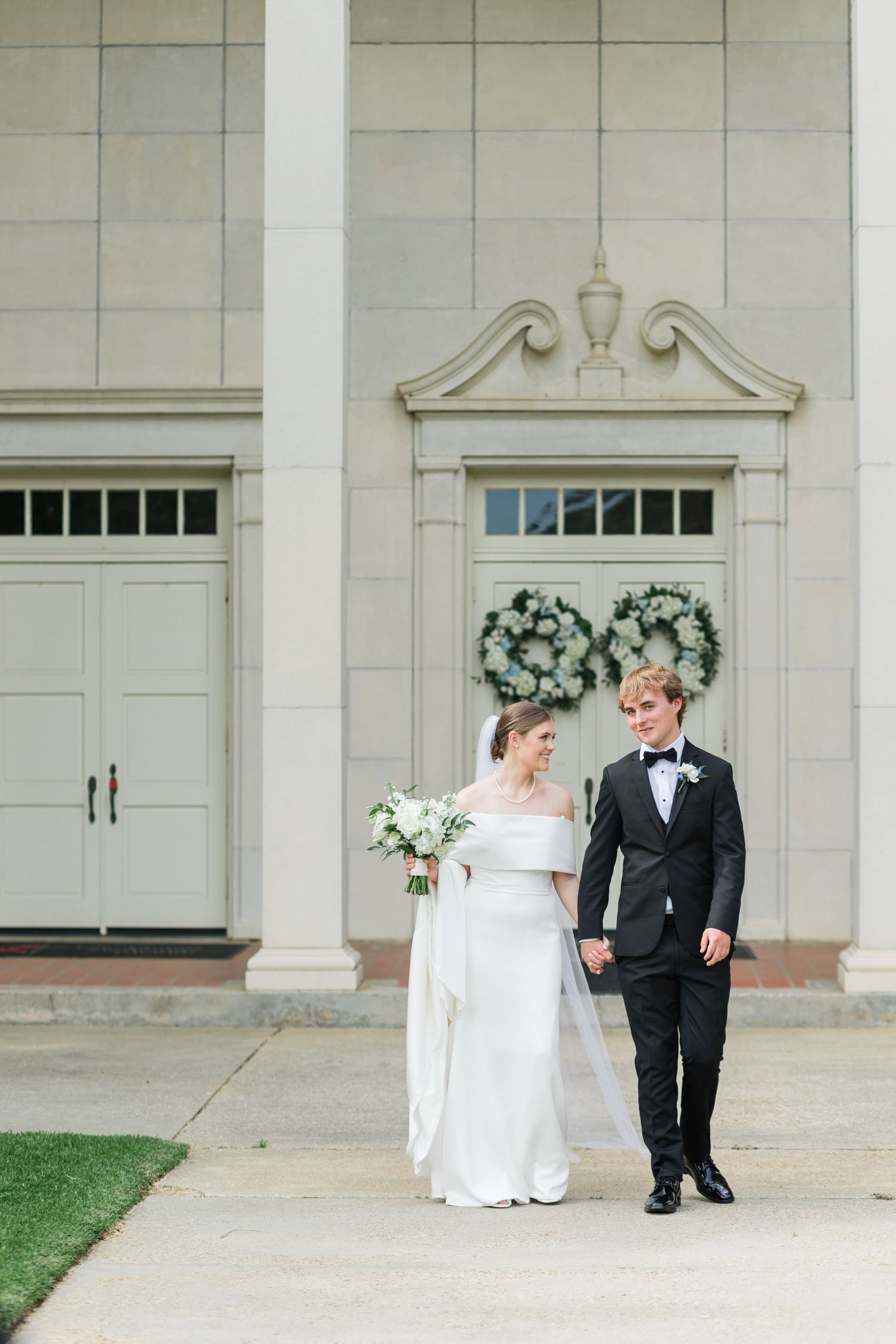 Bride and groom walking hand in hand in front of elegant building with wreath decorations.