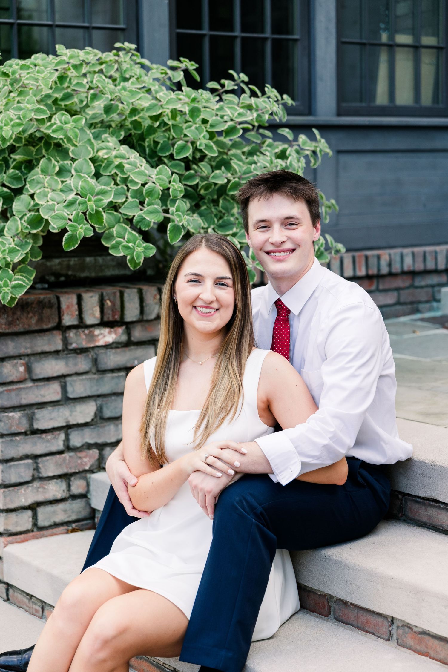 A smiling young couple sits on stone steps in front of a brick building surrounded by lush greenery.