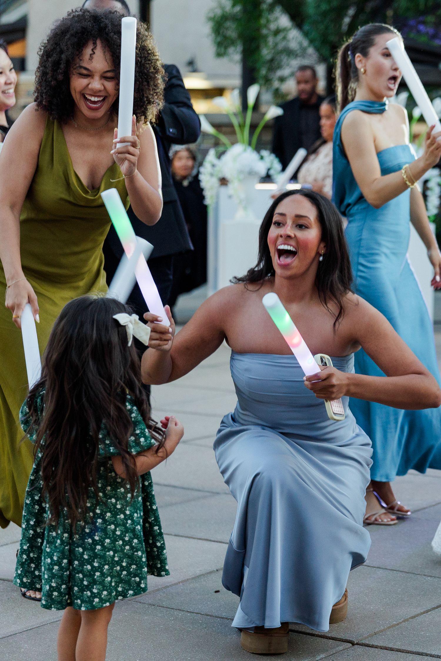 Women in formal dresses joyfully dancing with a little girl at an outdoor wedding celebration with glow sticks.