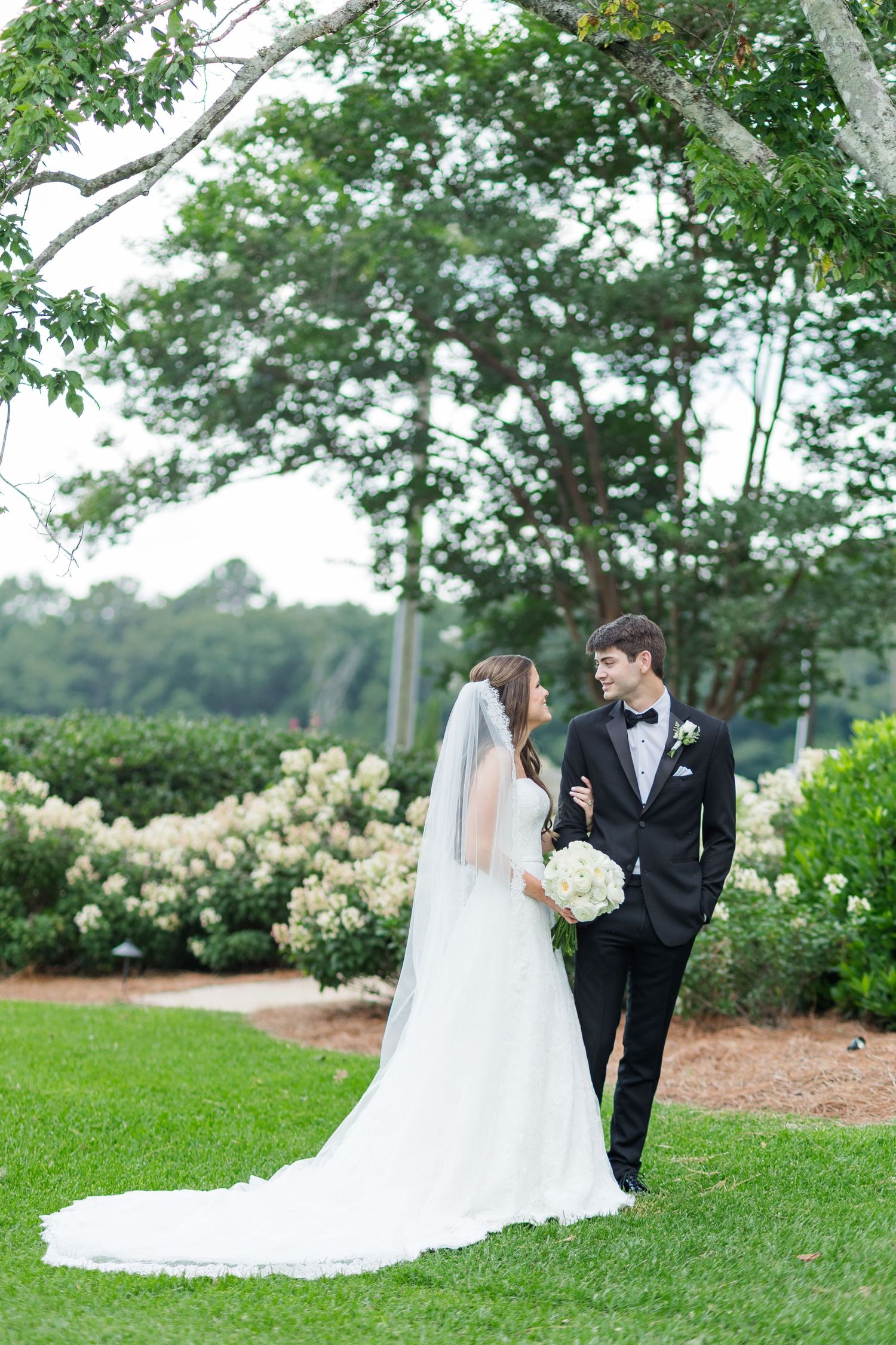 Bride and groom share a romantic moment on green lawn surrounded by white blooming flowers.