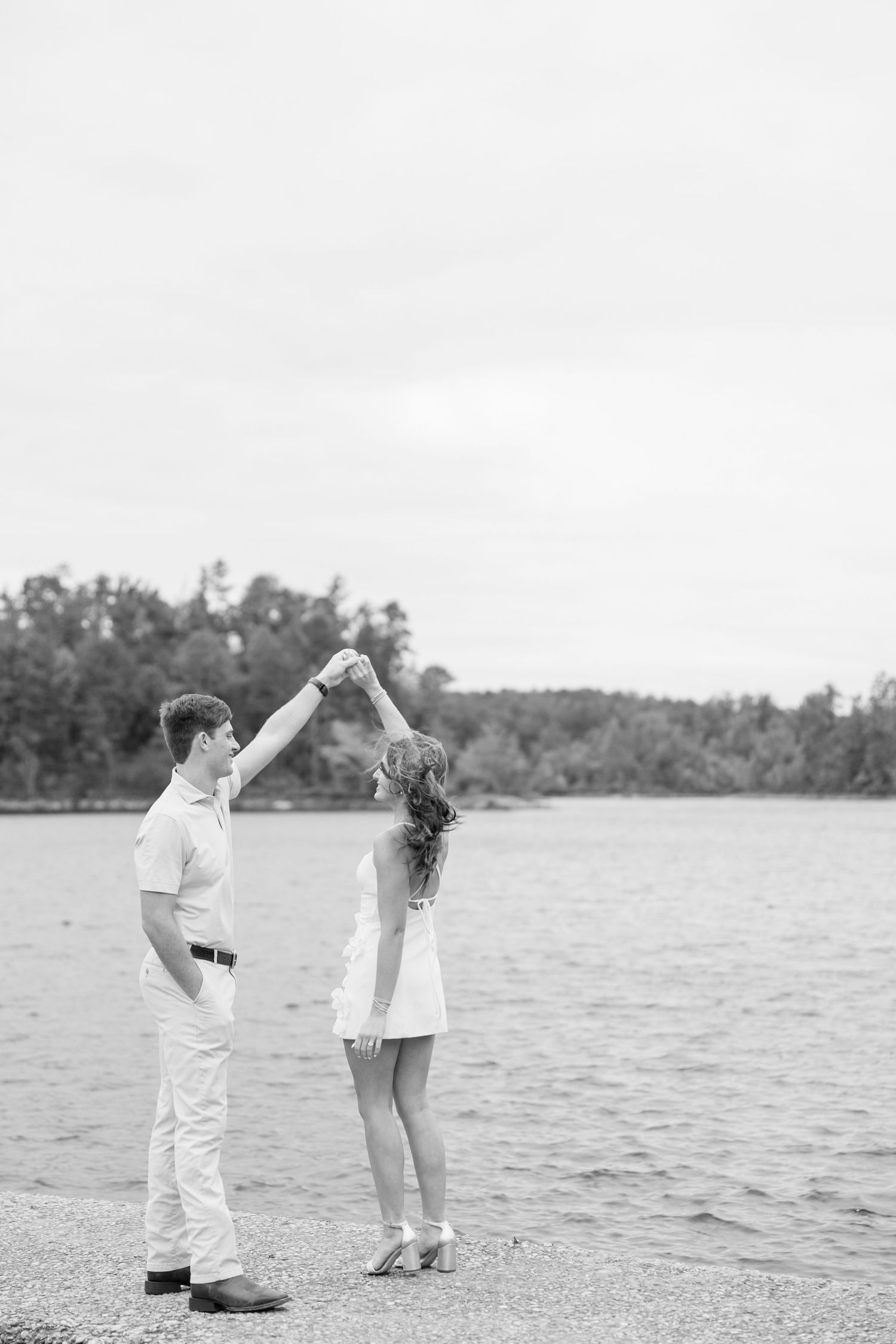 Black and white photo of a couple dancing by the lake shore, the man twirling the woman under his raised arm with trees in the distance.