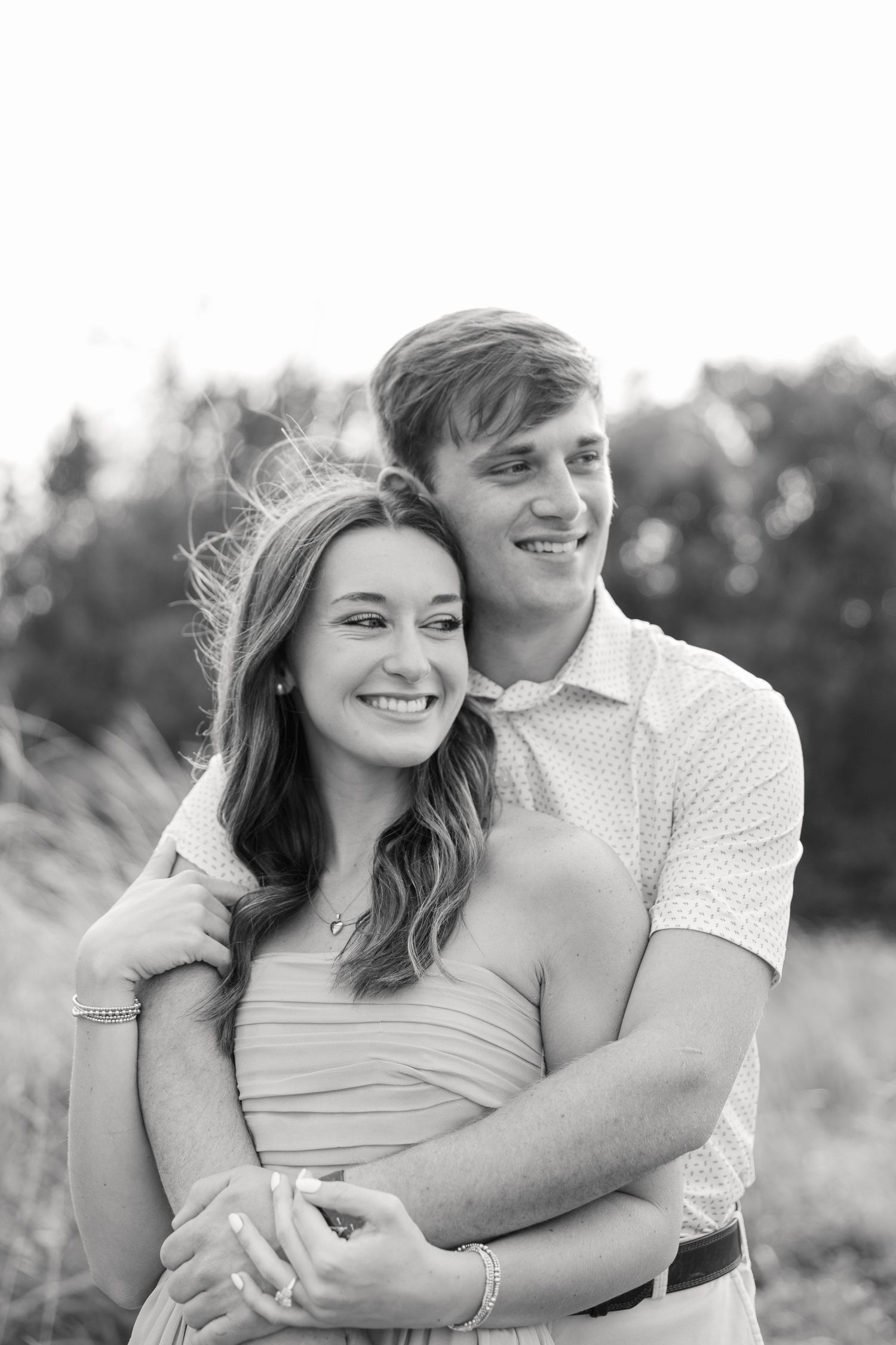 Black and white engagement portrait of a smiling couple embracing in a grassy field with trees softly blurred in the background.
