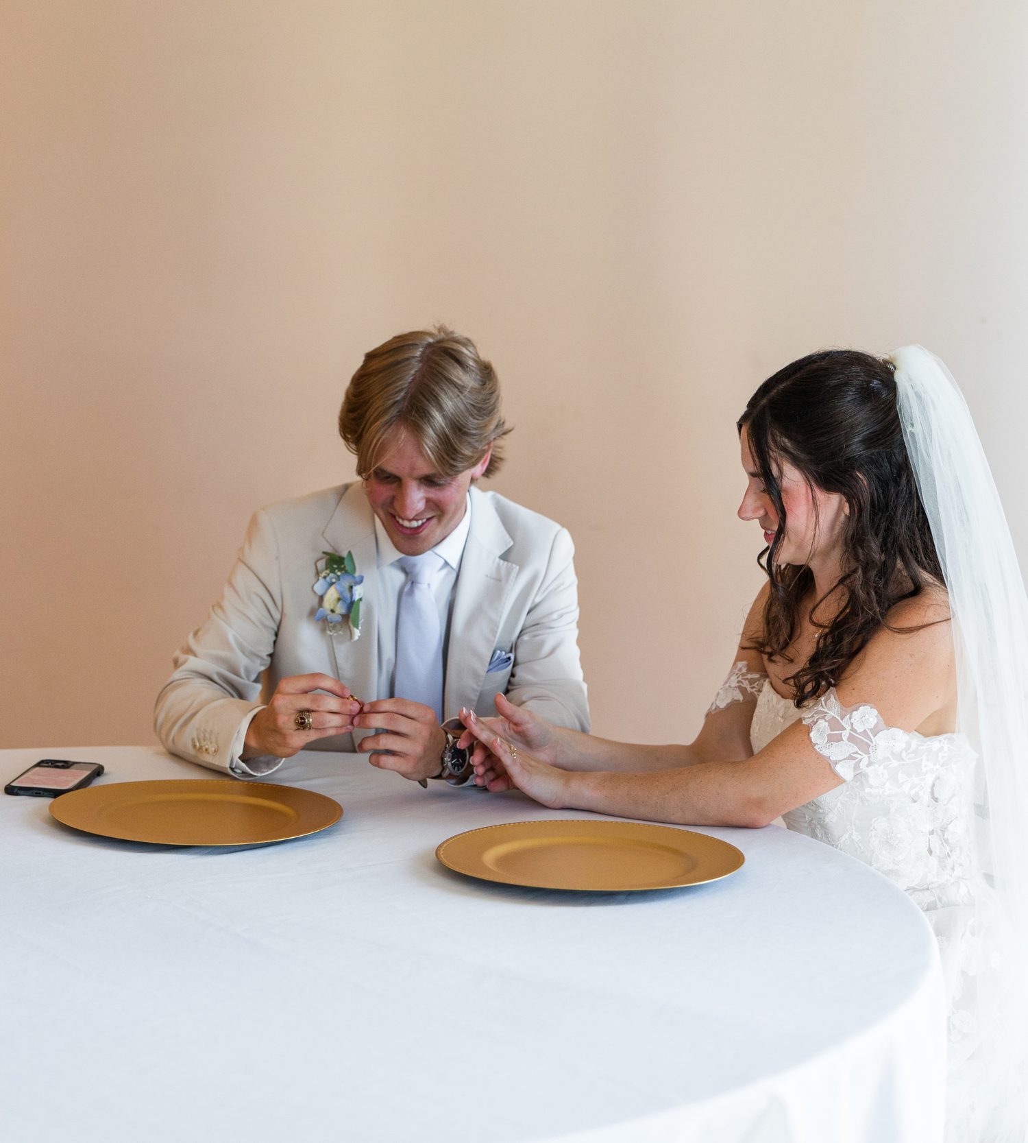 Bride and groom sitting at a white table with gold charger plates, exchanging rings and smiling at their wedding reception.