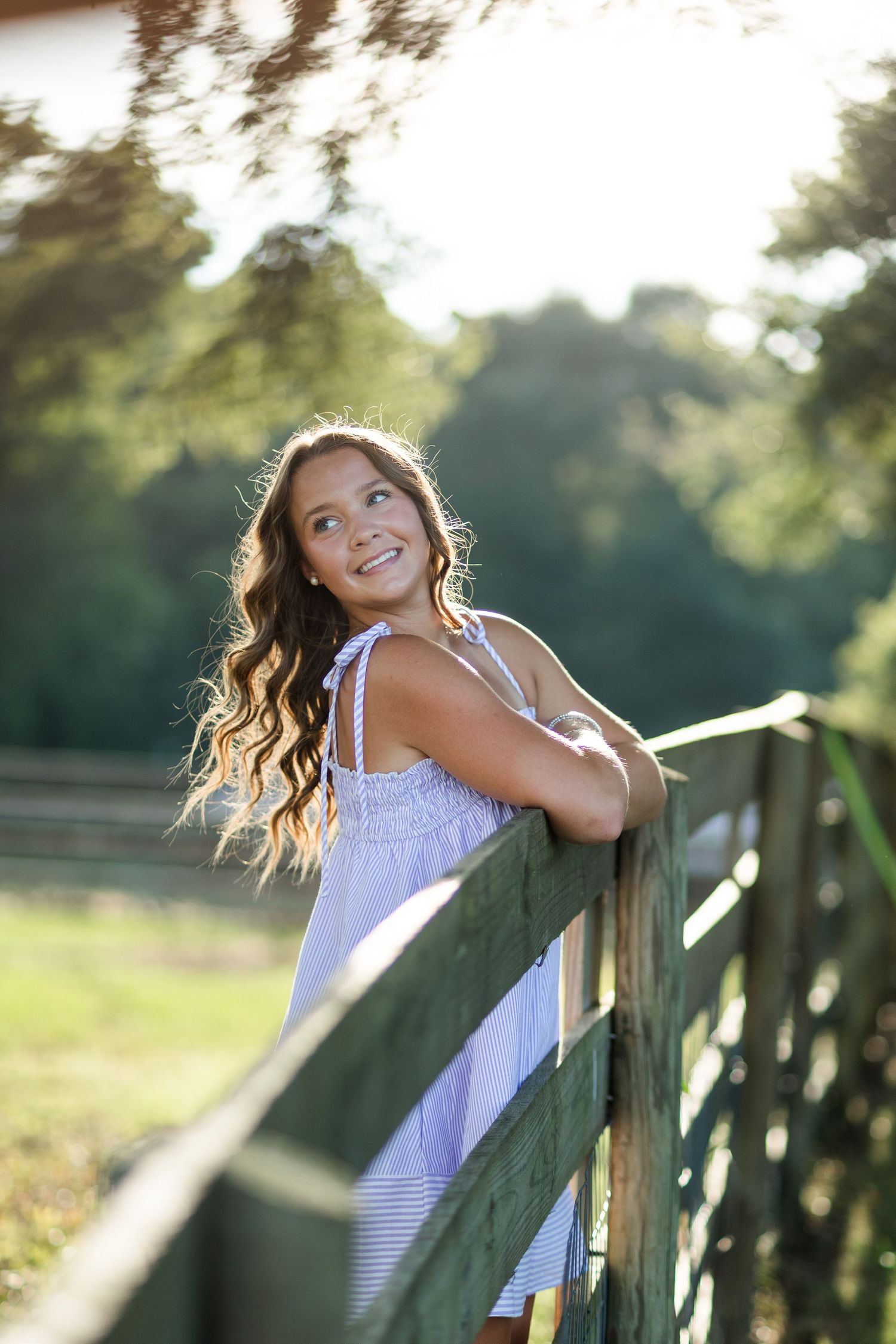 Young woman in blue dress leaning on wooden fence, smiling outdoors in golden sunlight with lush green trees.