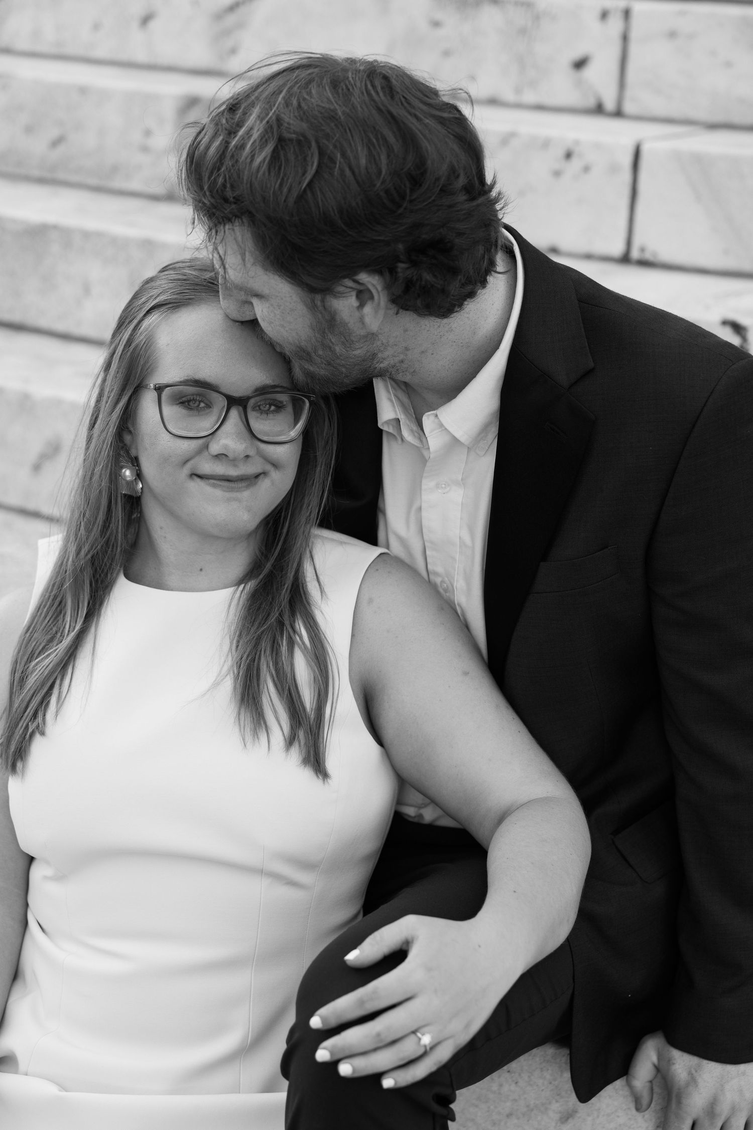 Black and white photo of smiling woman with glasses in white dress, man in suit kissing her head, engagement ring visible.