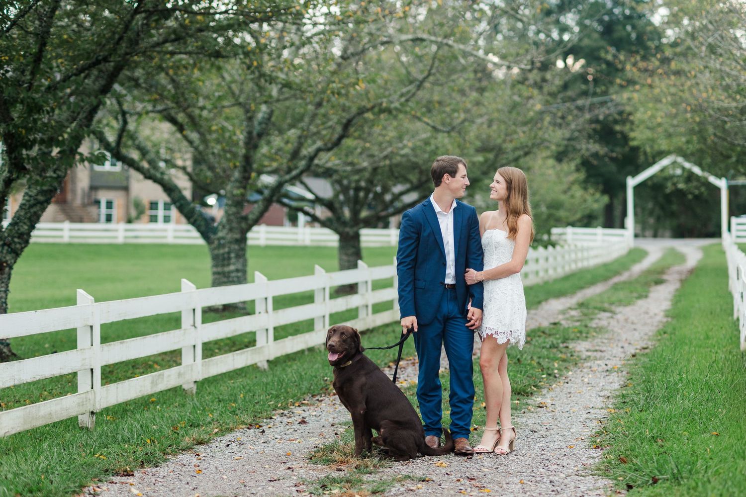 Couple stands on gravel path with chocolate Labrador, white fence, and green trees at a farm engagement session.
