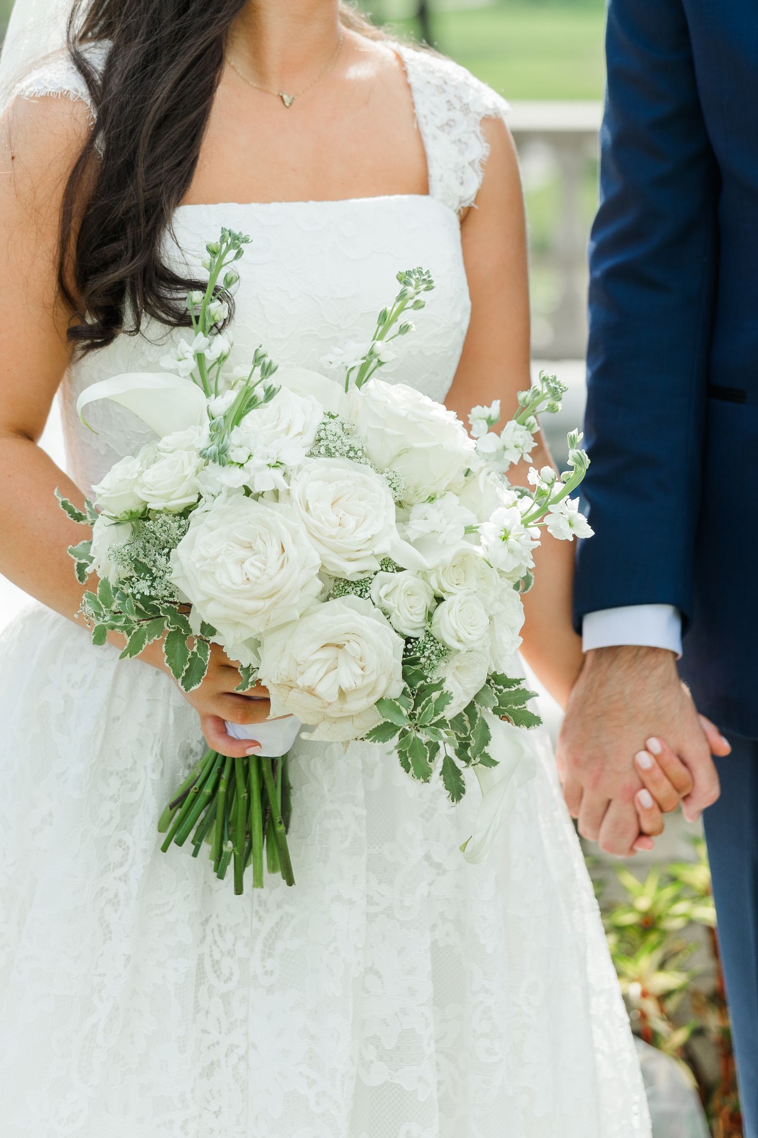 Bride holding a classic white rose and greenery wedding bouquet while holding grooms hand.