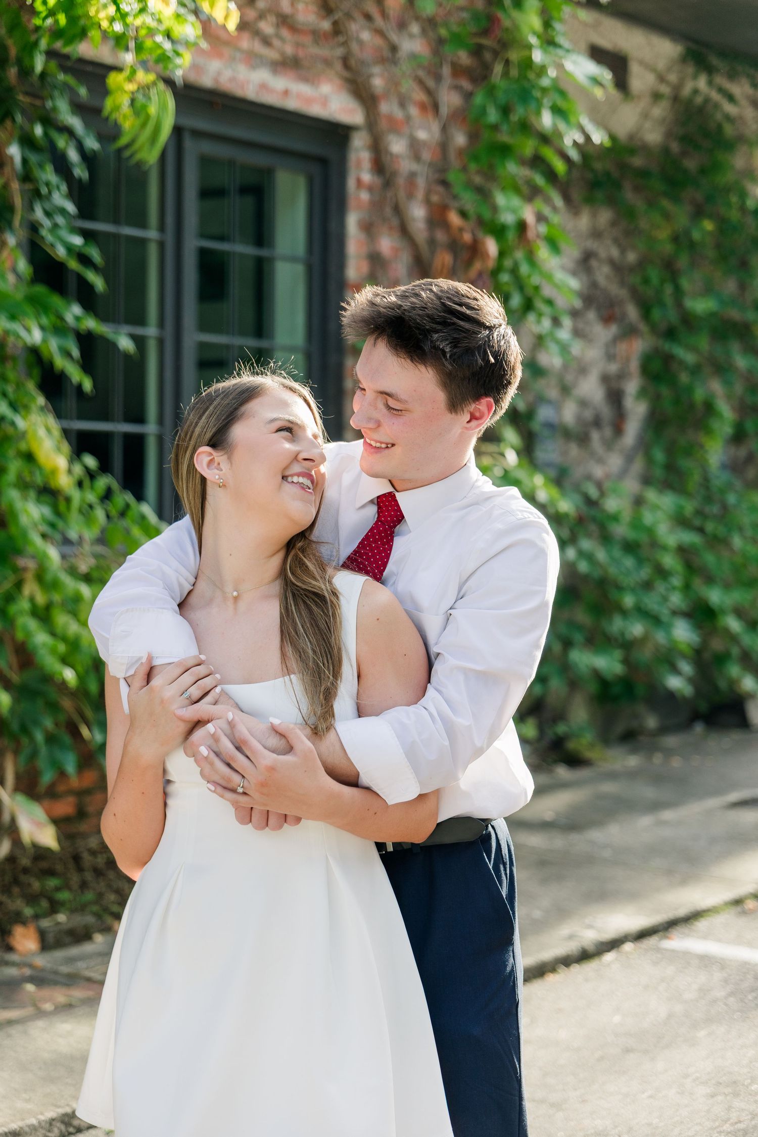 A couple embraces and smiles at each other outdoors, the man in a white shirt and red tie, the woman in a white dress.