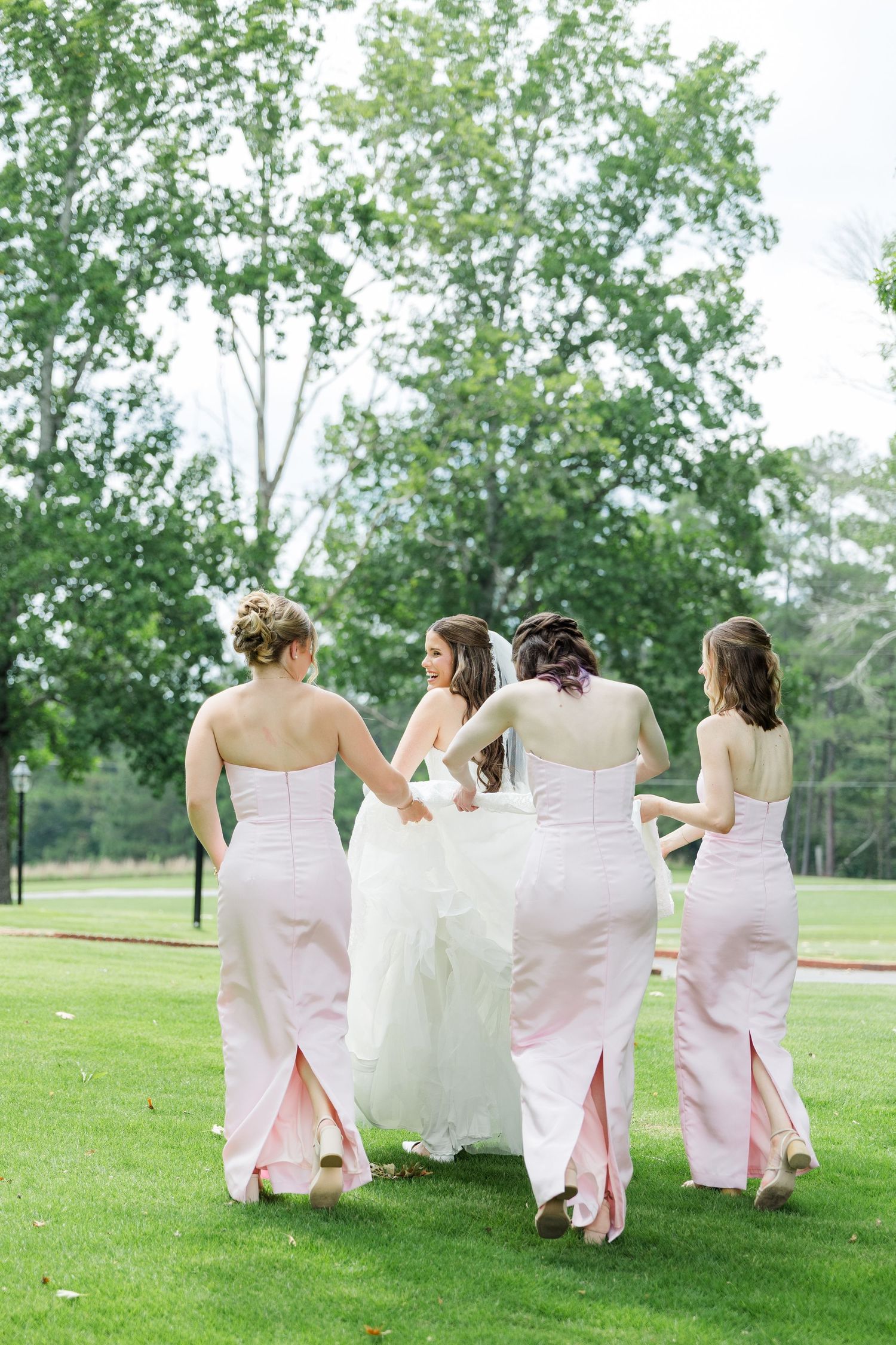 Bride in white gown walks with three bridesmaids in pink dresses across green lawn at outdoor wedding.