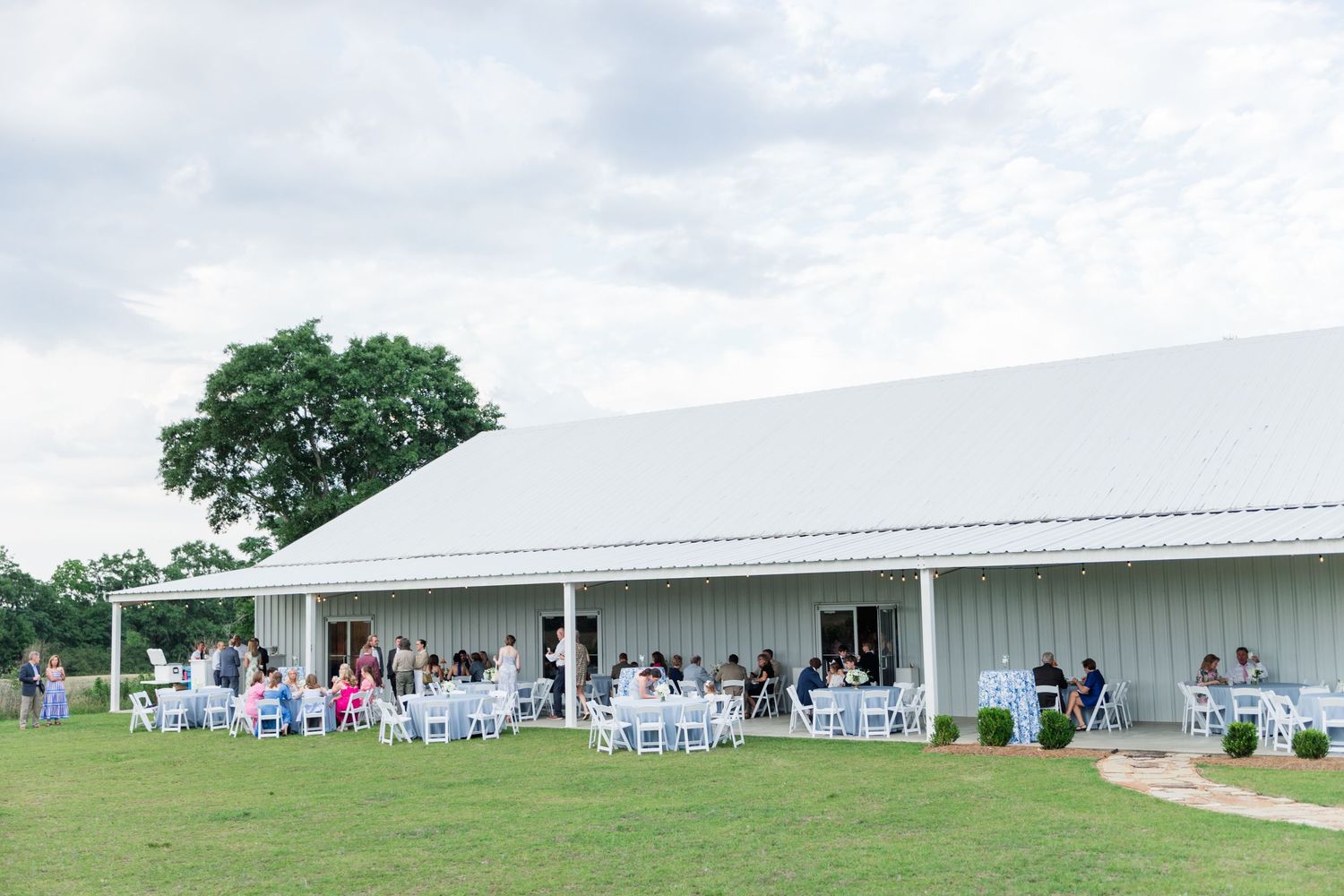 Outdoor wedding reception at a white barn venue with guests seated at round tables on a lush green lawn under cloudy skies.