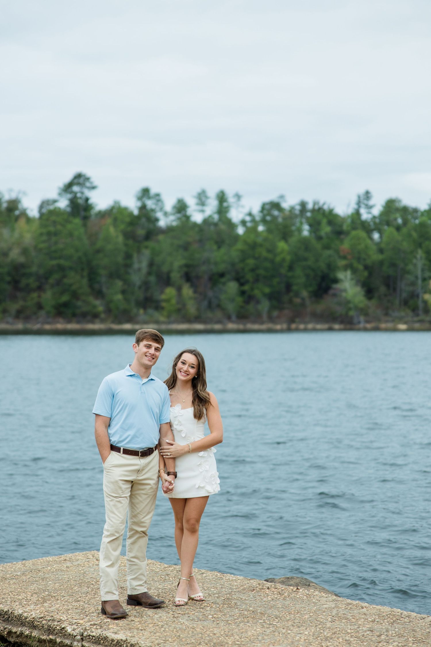 Smiling couple in a blue polo and white dress stand arm in arm on a stone pier overlooking a calm lake and tree-lined shore.