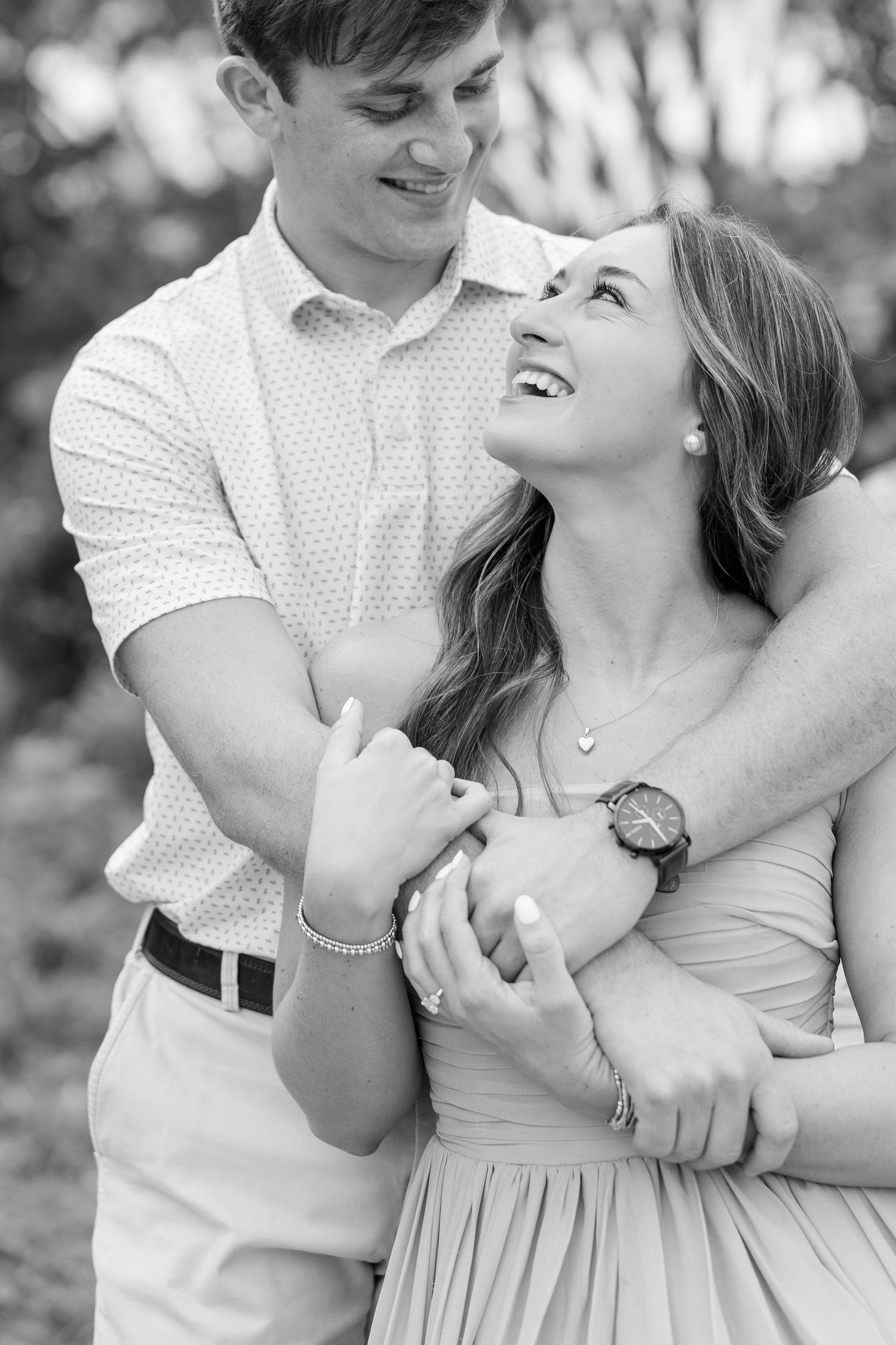 Black and white engagement photo of a smiling couple embracing outdoors as the woman gazes up at the man while he hugs her from behind.