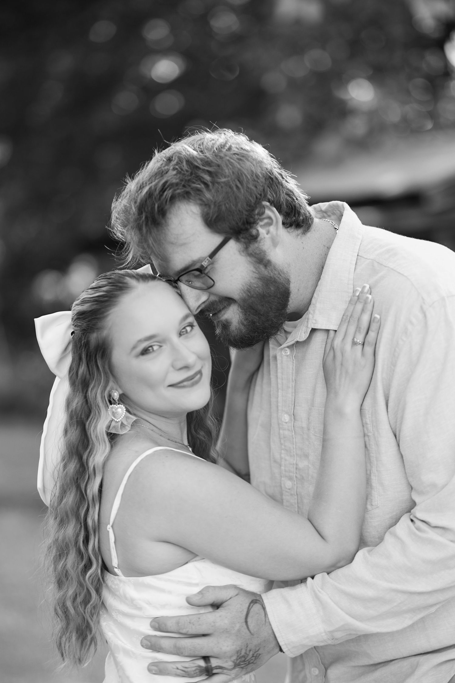 Black and white portrait of couple embracing, woman in white dress smiling while bearded man with glasses holds her close.