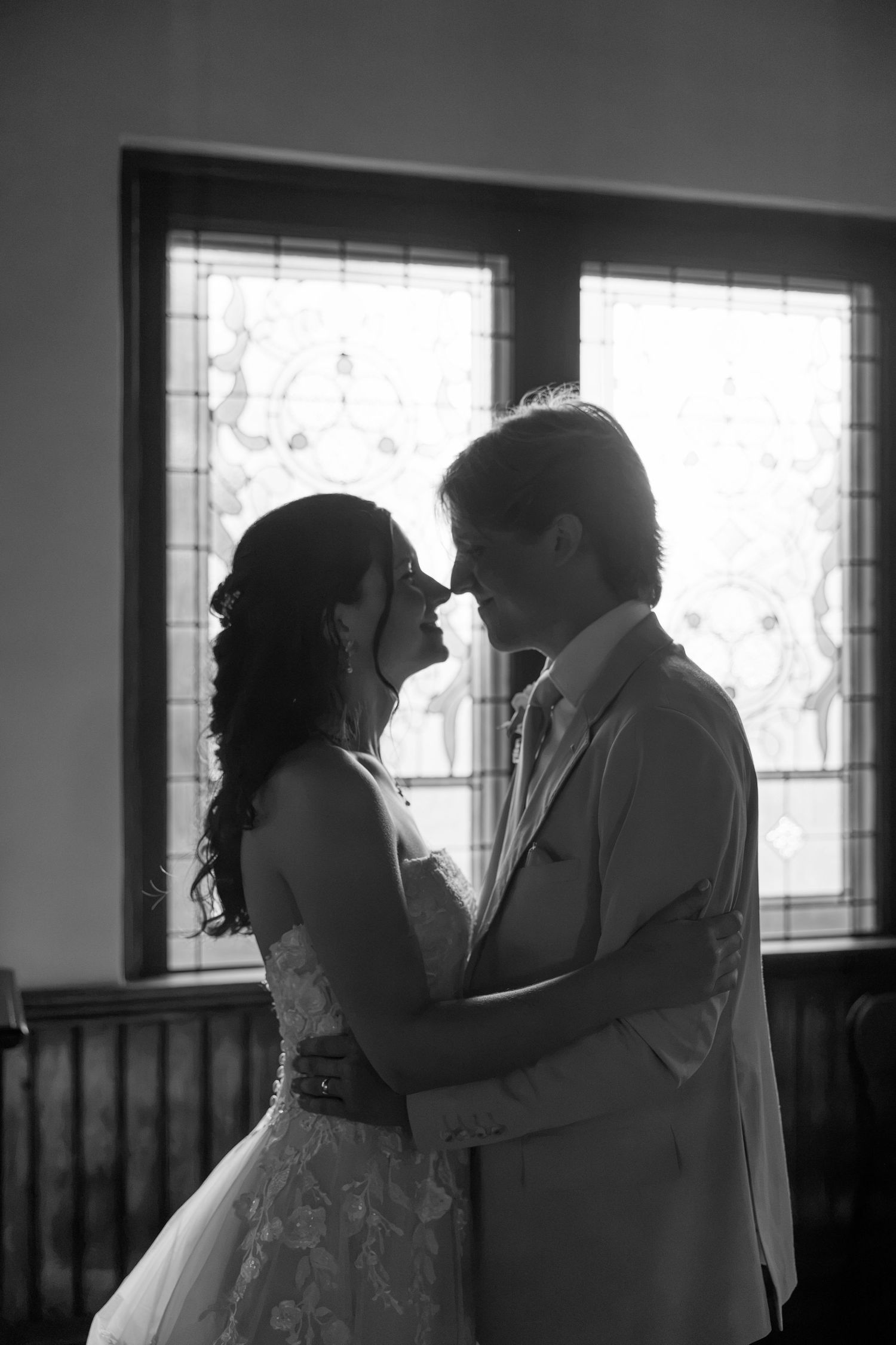 Black and white photo of bride and groom sharing romantic moment in front of stained glass church window.