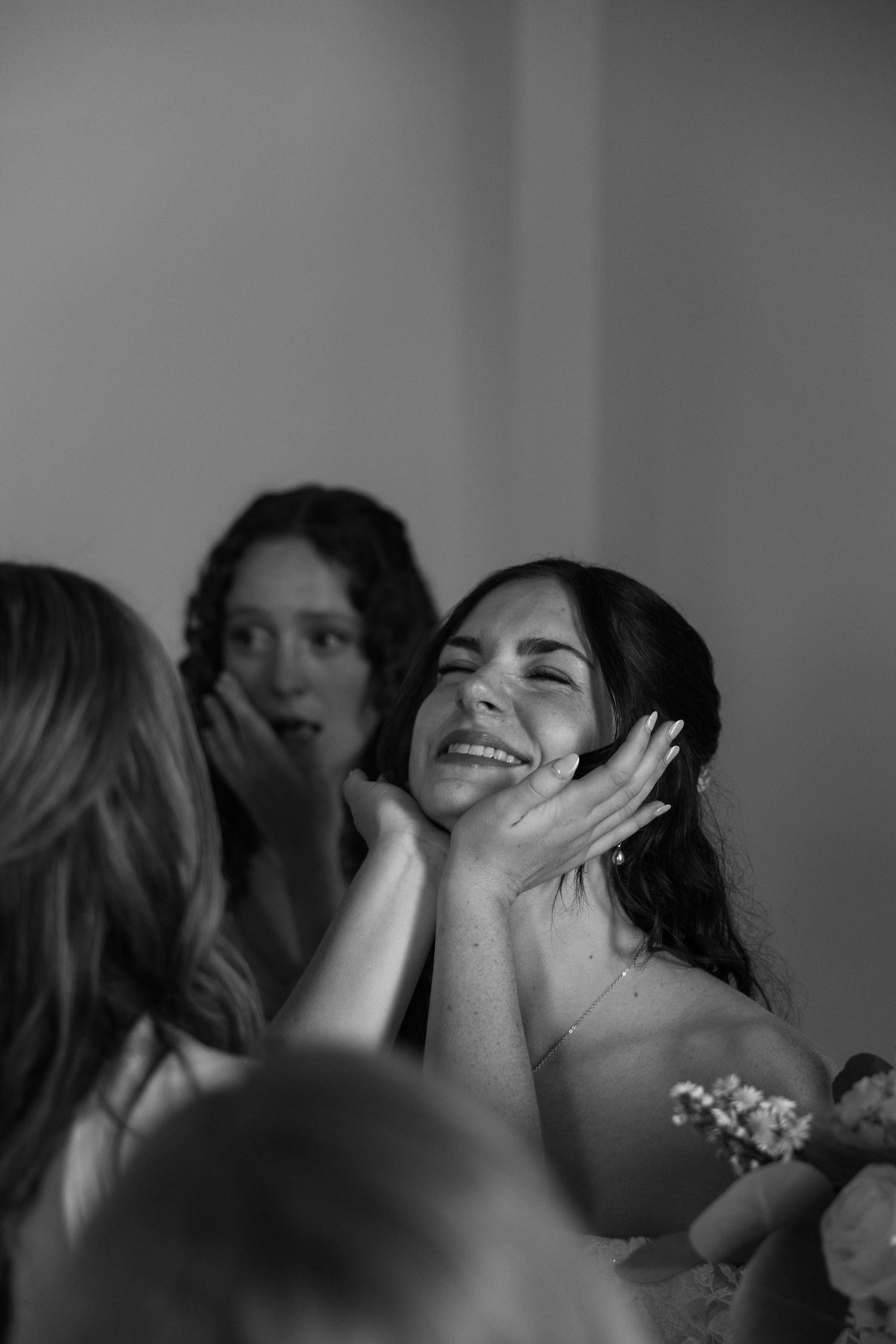 Black and white photo of a smiling bride having her face held by loved ones during wedding preparations.