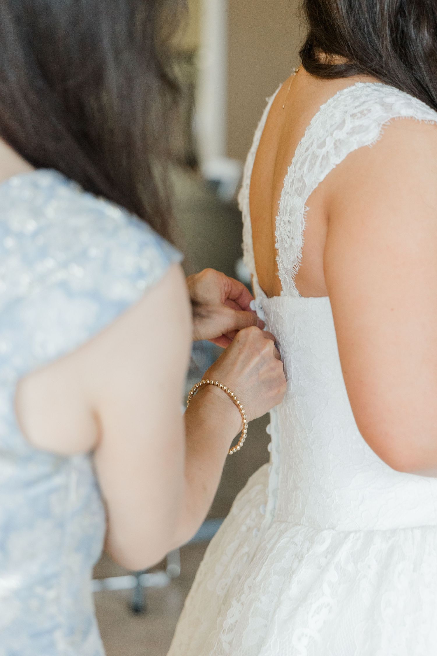 A bridesmaid in a blue dress fastens the back buttons of a bride's white lace wedding gown.