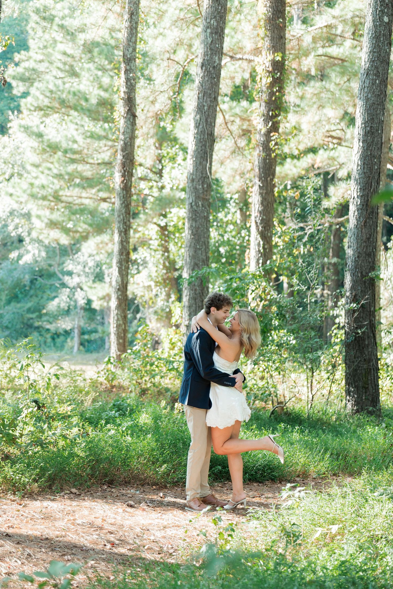 Couple sharing a romantic kiss in a sunlit forest, surrounded by tall pine trees and lush green foliage.