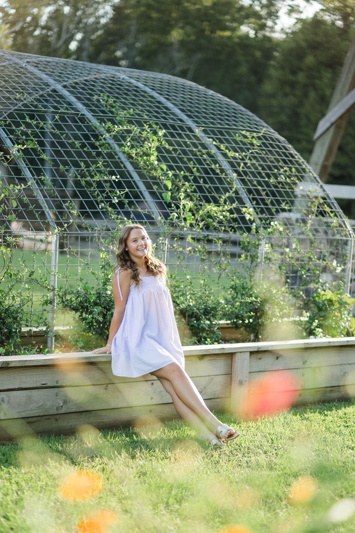 Young woman in a white sundress sitting on a wooden raised garden bed in front of a dome trellis.
