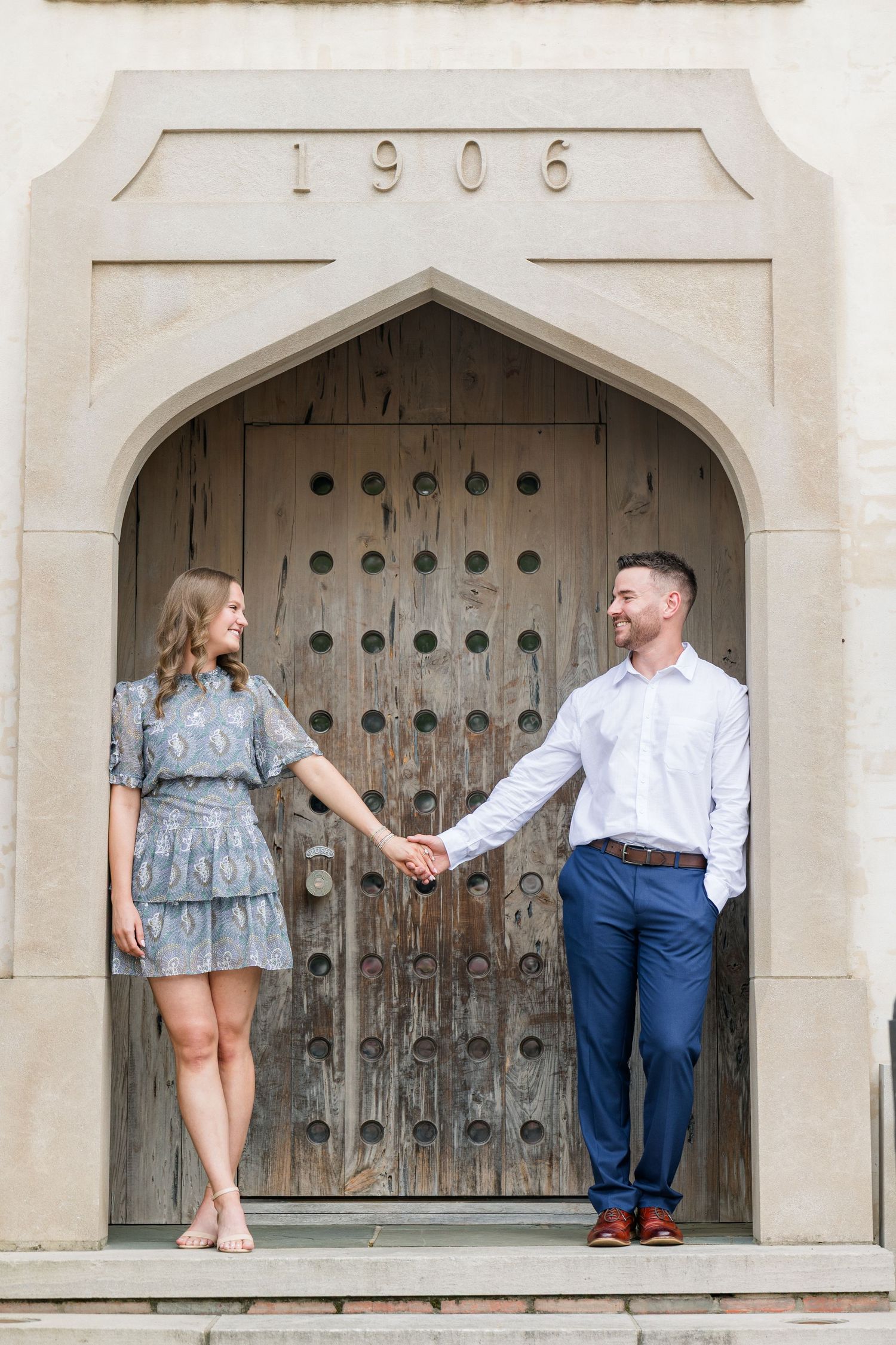 Couple holding hands in front of a historic 1906 arched wooden door, dressed in elegant blue outfits.