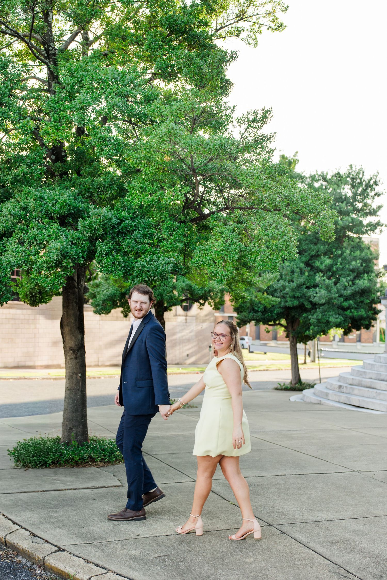 Couple holding hands walking outdoors, man in navy suit, woman in yellow dress, green trees in background.