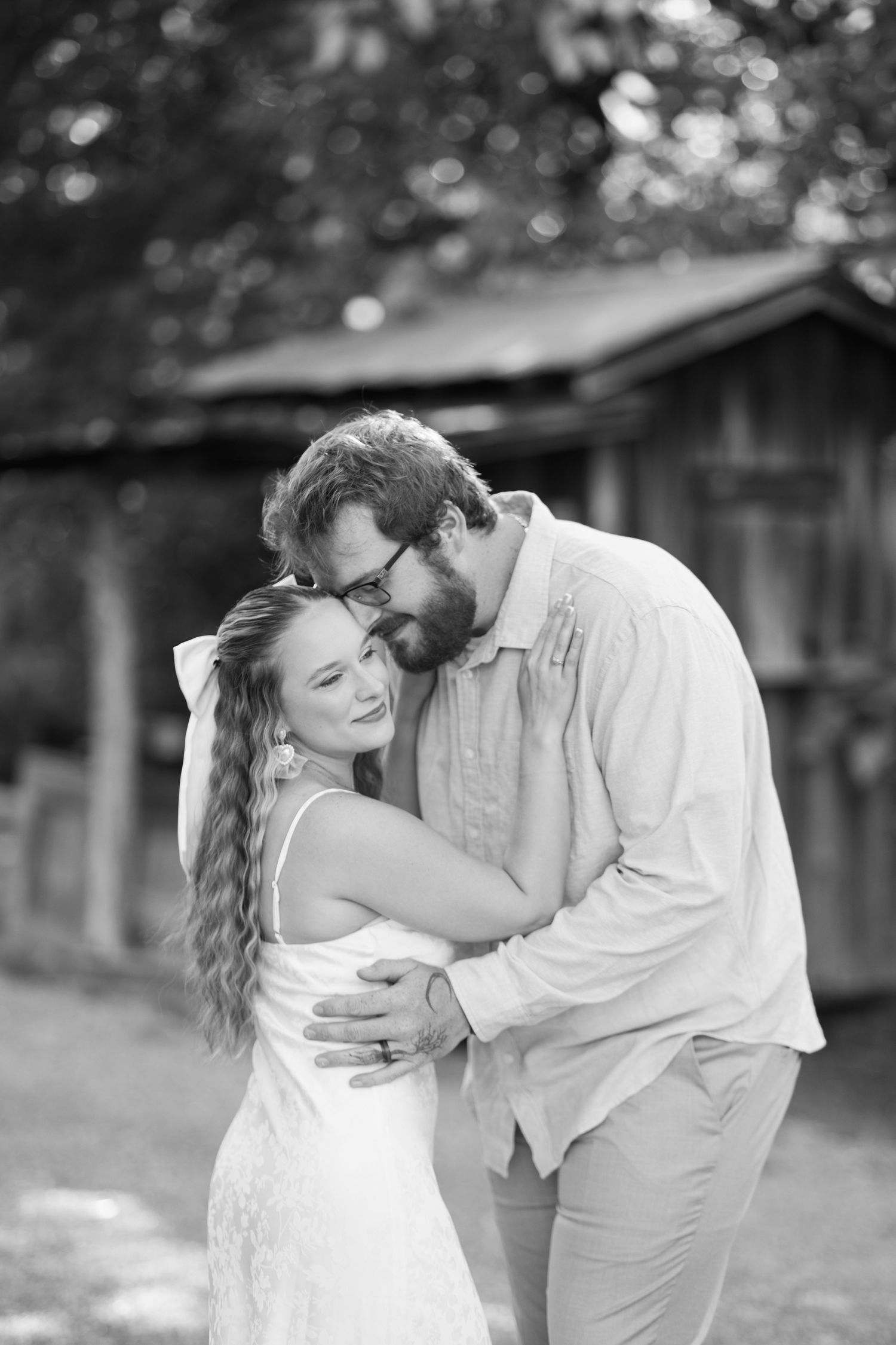 Black and white photo of a couple embracing outdoors, woman in white dress smiling as man with glasses kisses her head.