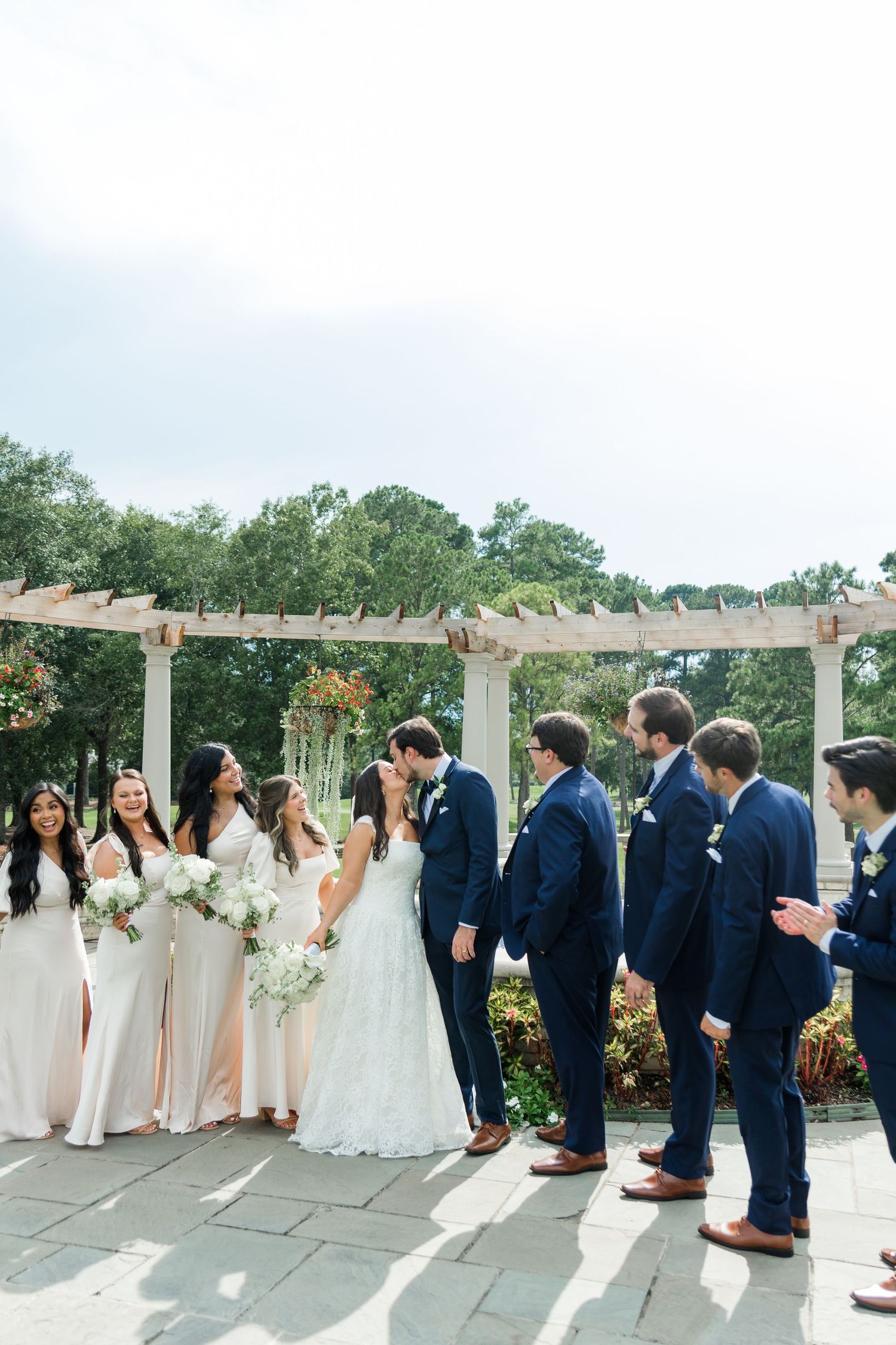 Wedding party in navy suits and white gowns celebrating with bride and groom kissing under pergola.