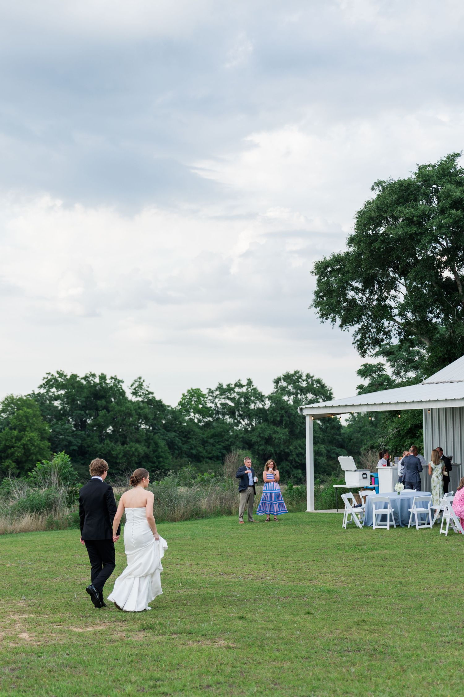 A bride and groom walk hand-in-hand across a green lawn toward their outdoor wedding reception tent.