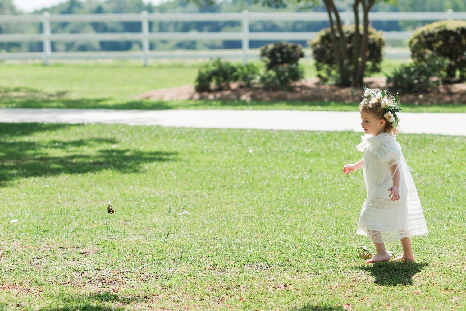 Adorable flower girl in white dress and floral crown runs across green lawn at outdoor wedding venue.