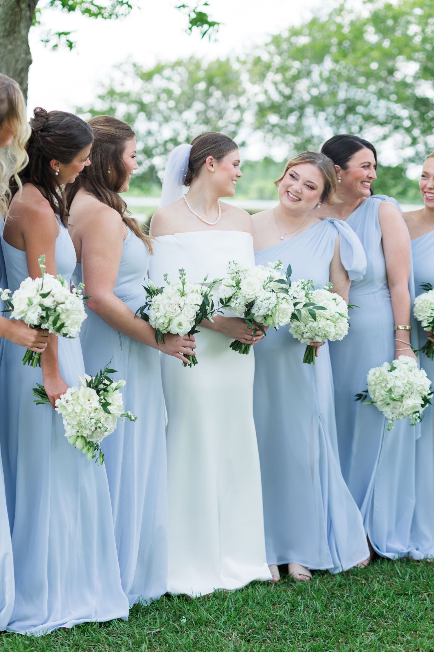 Bride in white gown with bridesmaids in light blue dresses holding white floral bouquets outdoors.