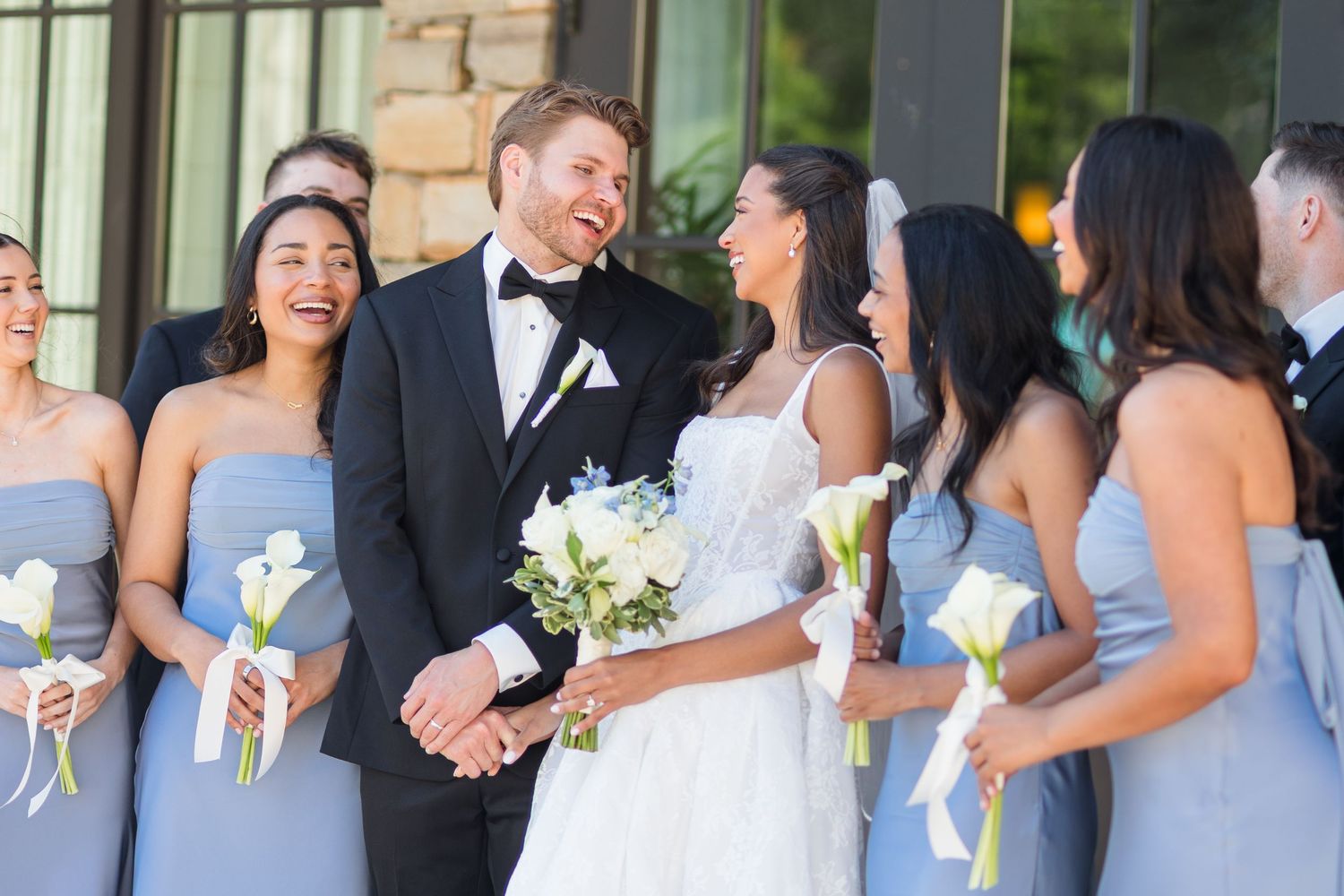Bride and groom laugh with bridesmaids in light blue strapless dresses holding white bouquets at outdoor wedding.