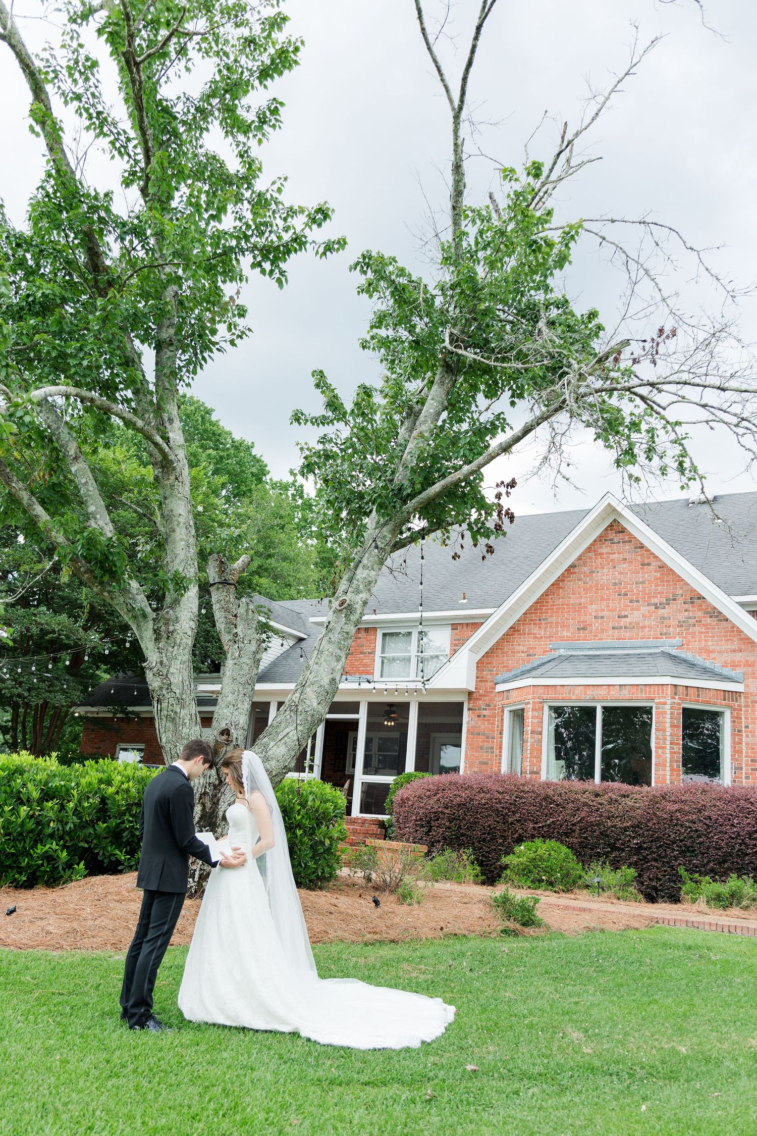 Bride and groom share a kiss under a tree outside a brick wedding venue.
