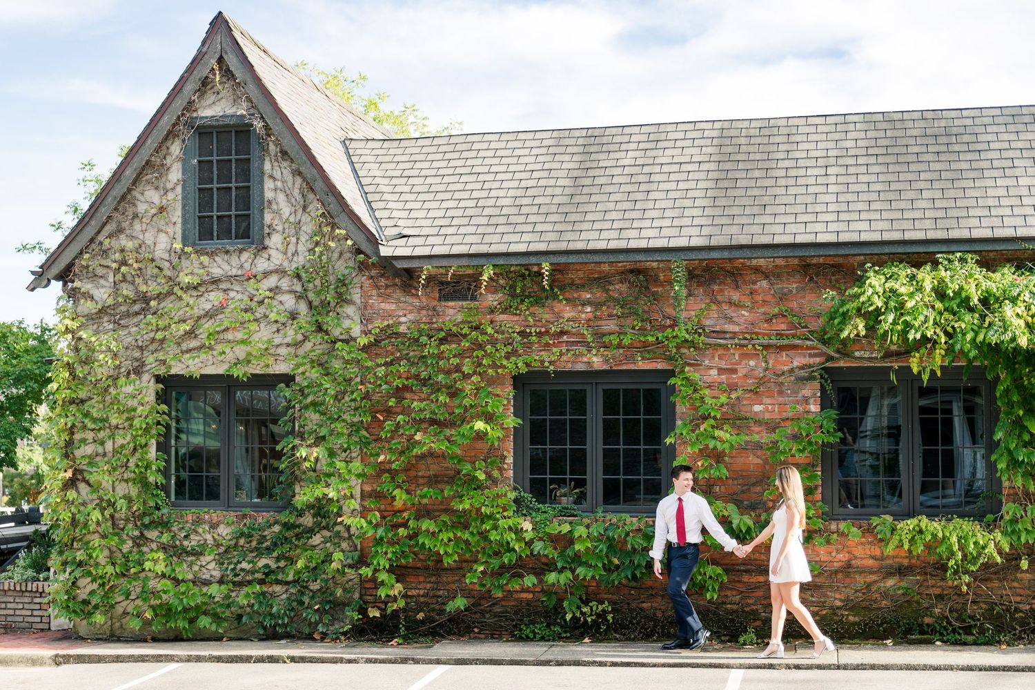 A couple holds hands walking past a charming ivy-covered brick building on a sunny day.