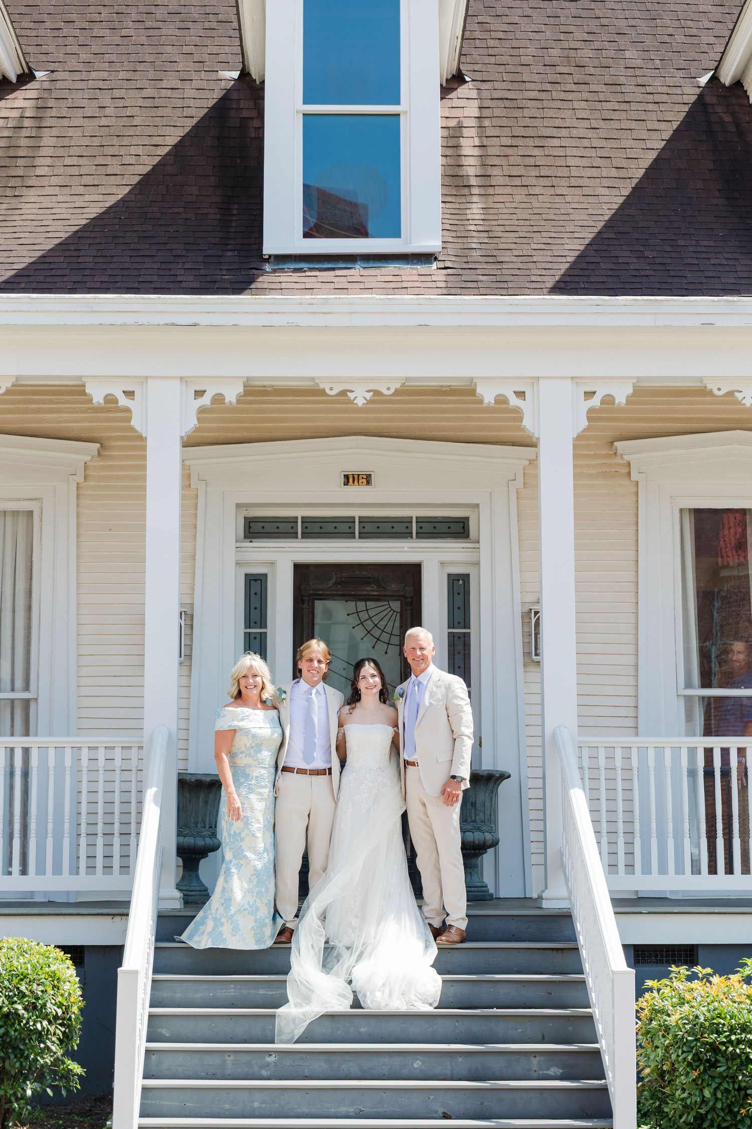 Wedding party of four poses on steps of white Southern colonial home with wraparound porch on sunny day.