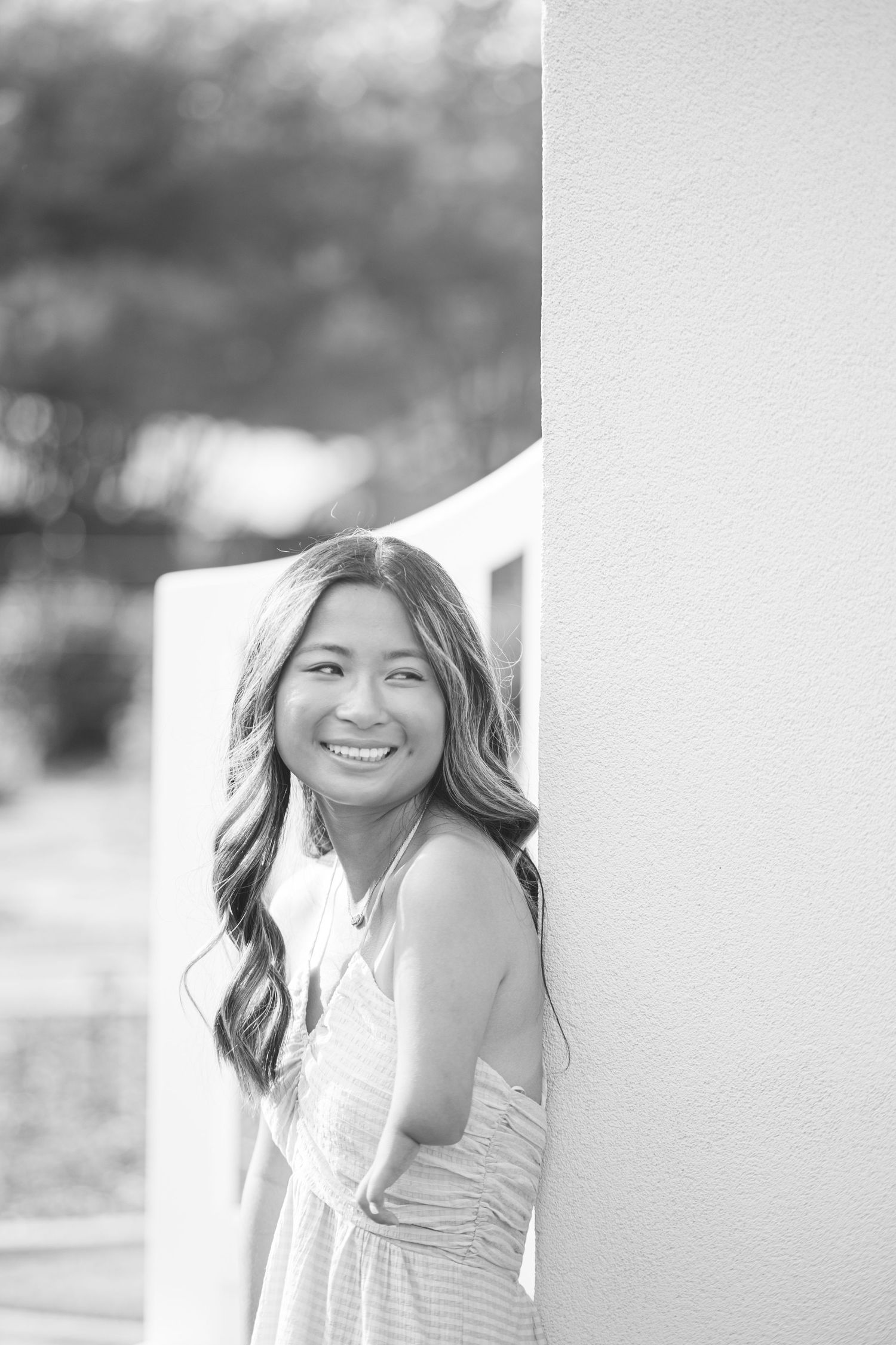 Young woman in sundress leaning against white column smiling in black and white portrait photo.