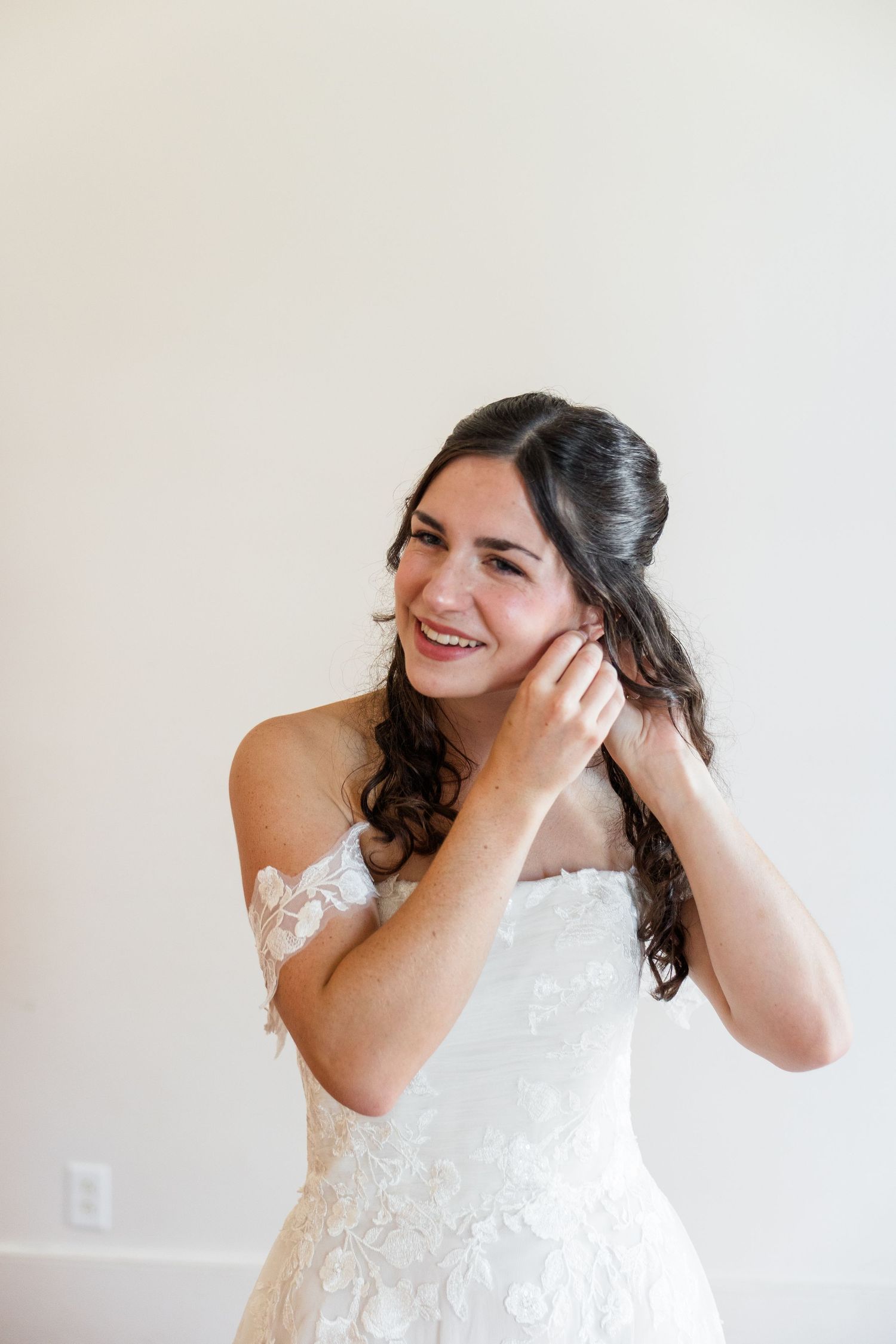 Smiling bride in off-shoulder lace wedding gown puts on earring while getting ready on her wedding day.