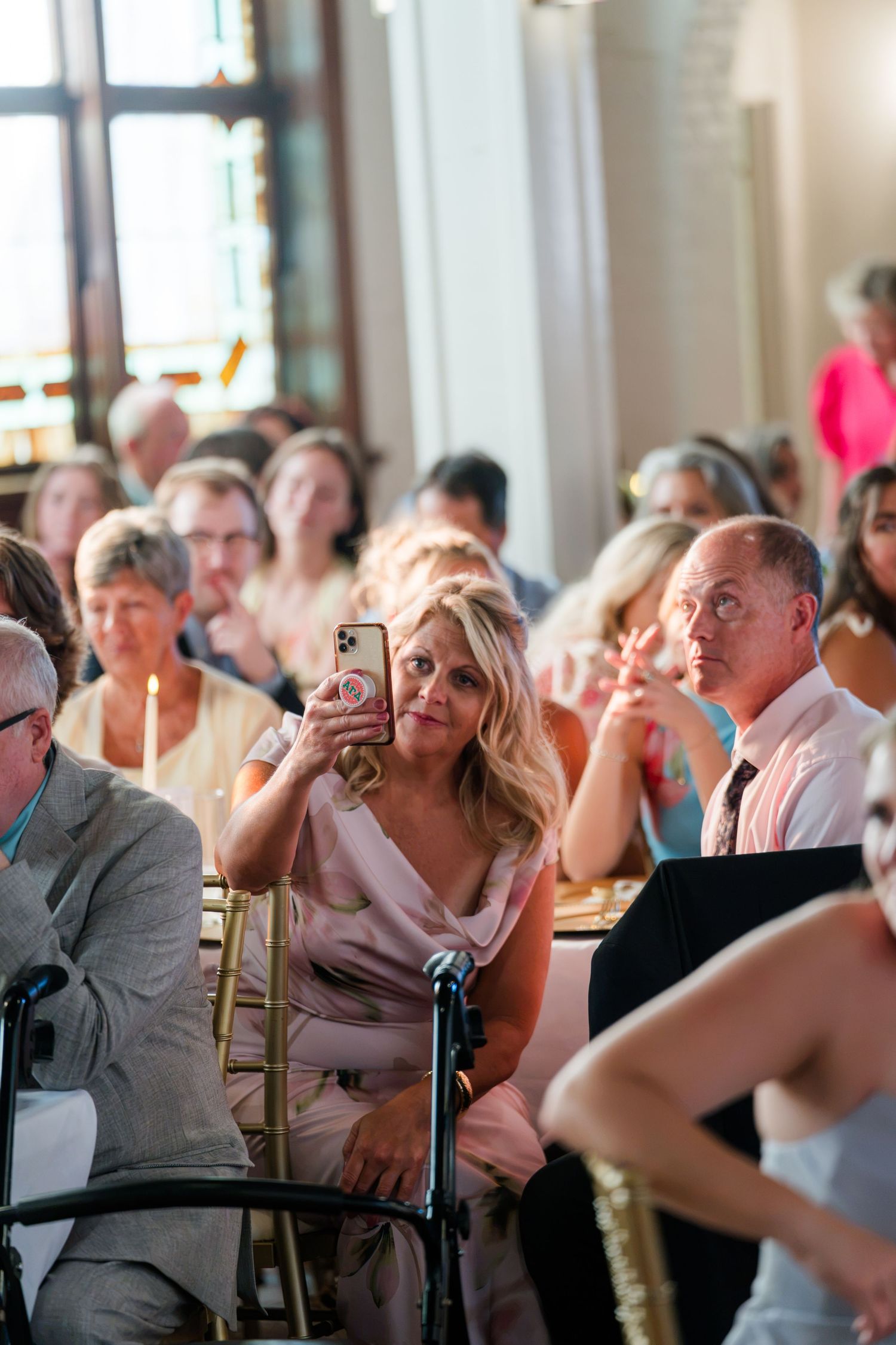 Wedding guests seated at a ceremony, a blonde woman photographs with her iPhone while others watch attentively.
