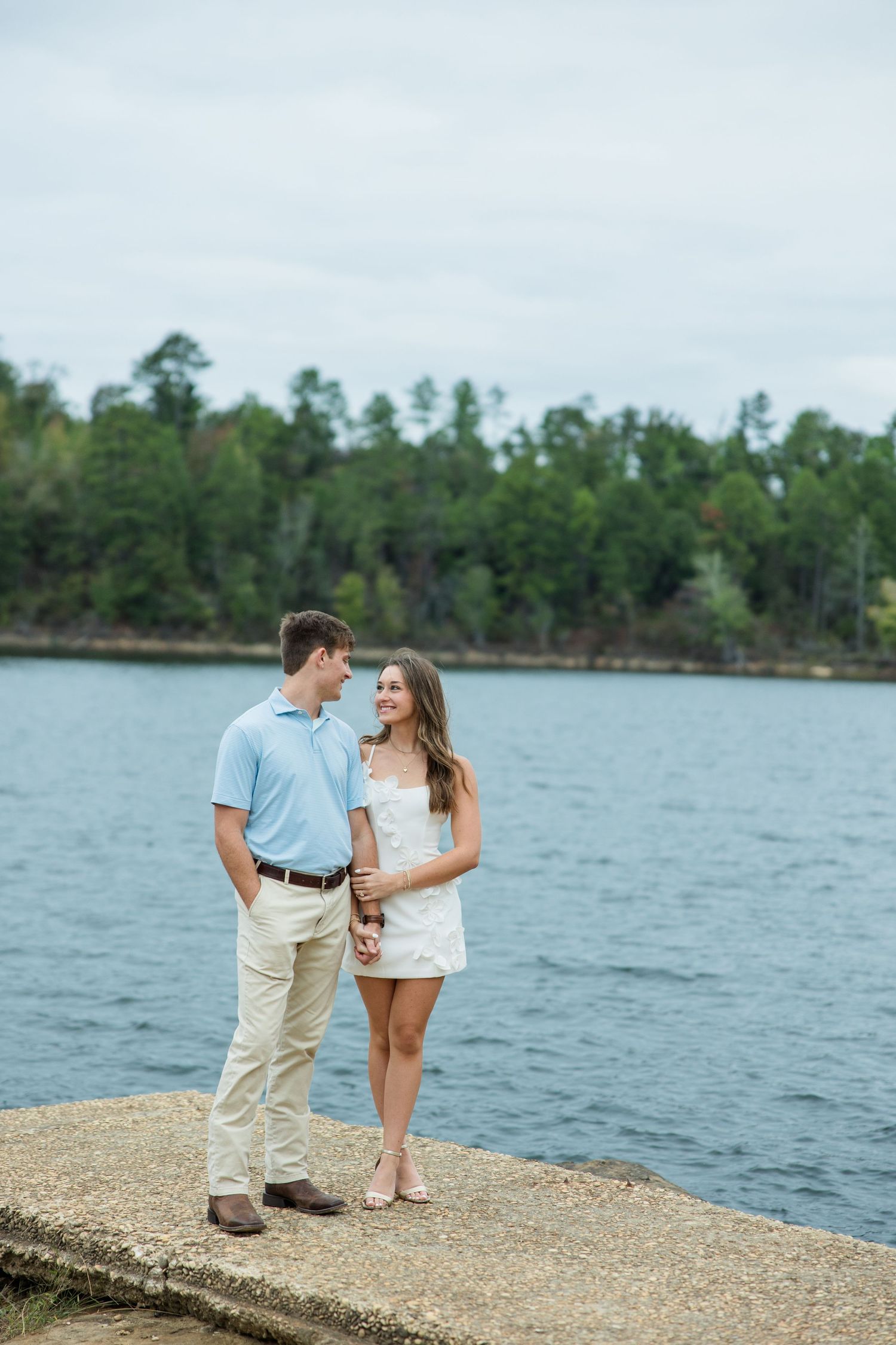 Couple in a light blue polo and white dress share a happy glance while standing on a concrete lakeside pier with forested shoreline behind.