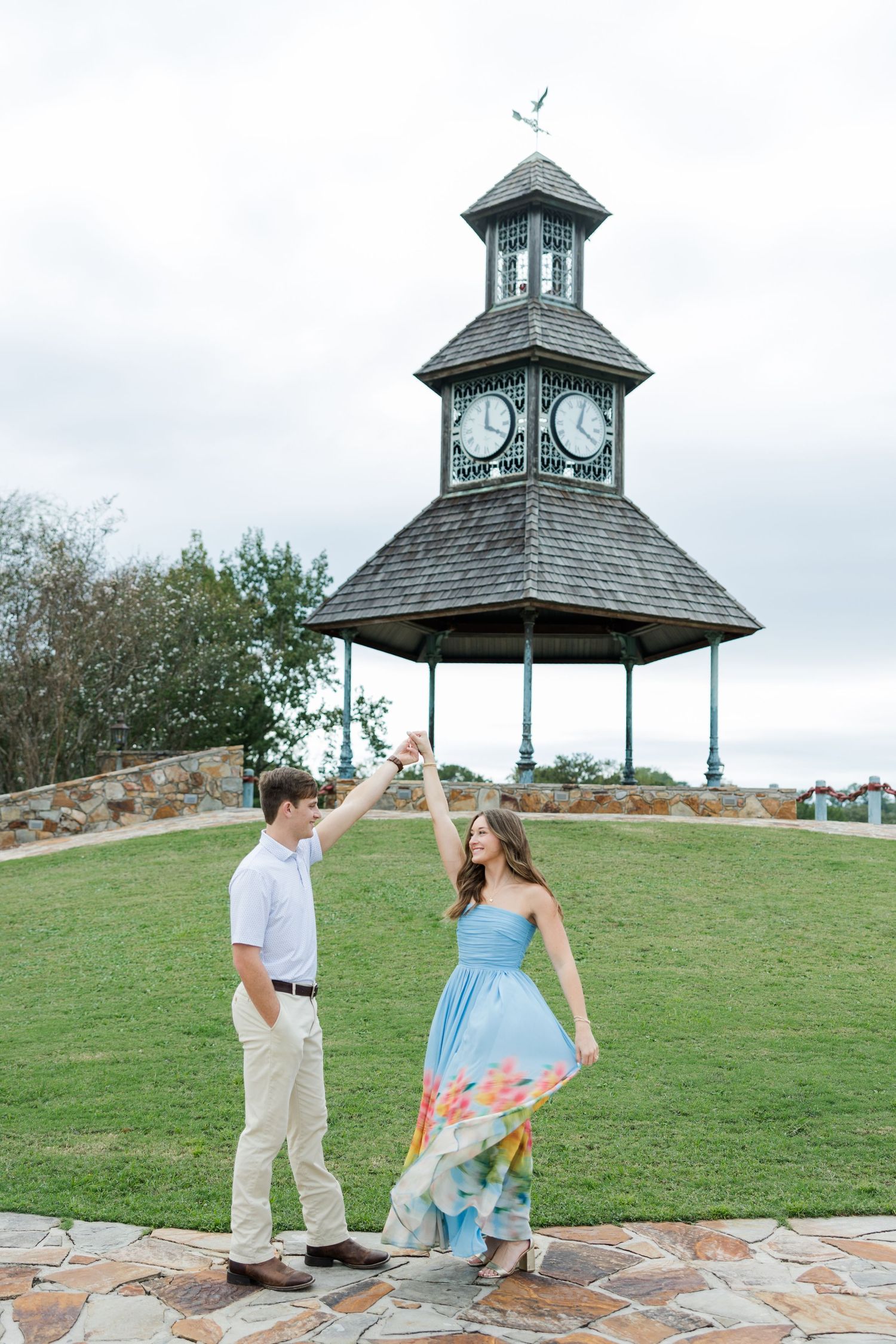 A couple dances hand in hand on a stone patio, her floral gown twirling, with a clock tower gazebo and green lawn behind them.