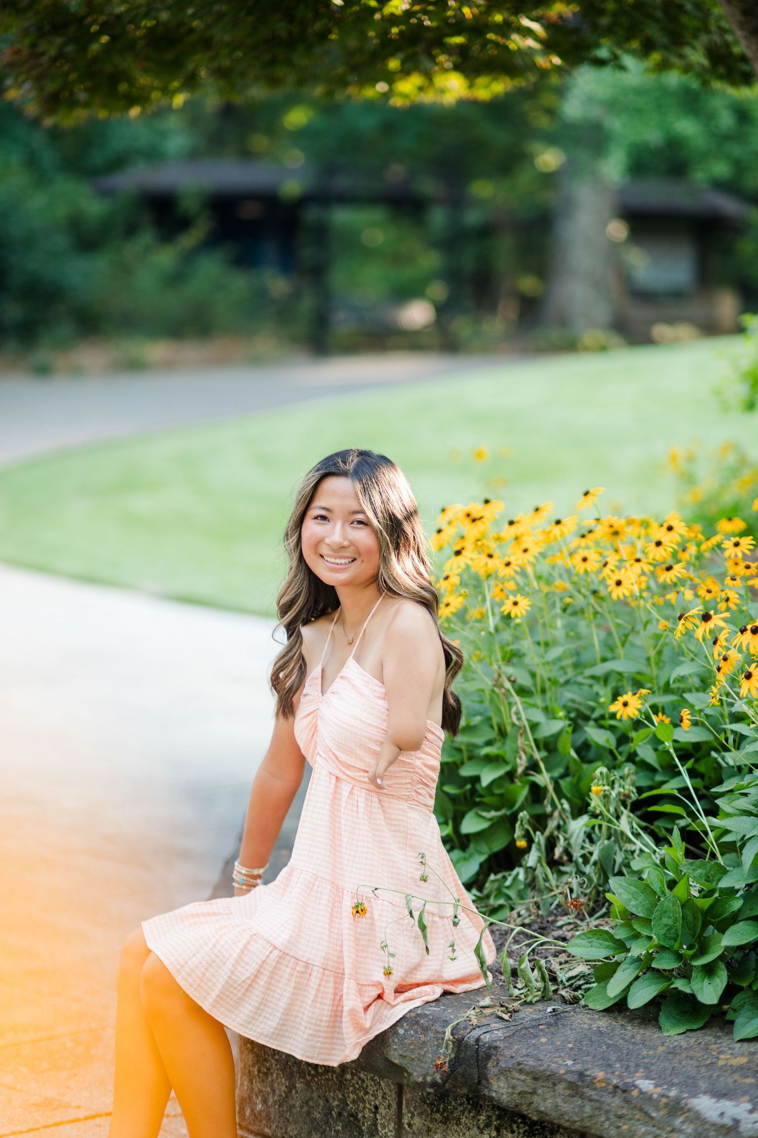 Young woman in peach floral dress sitting on stone wall beside yellow flowers.